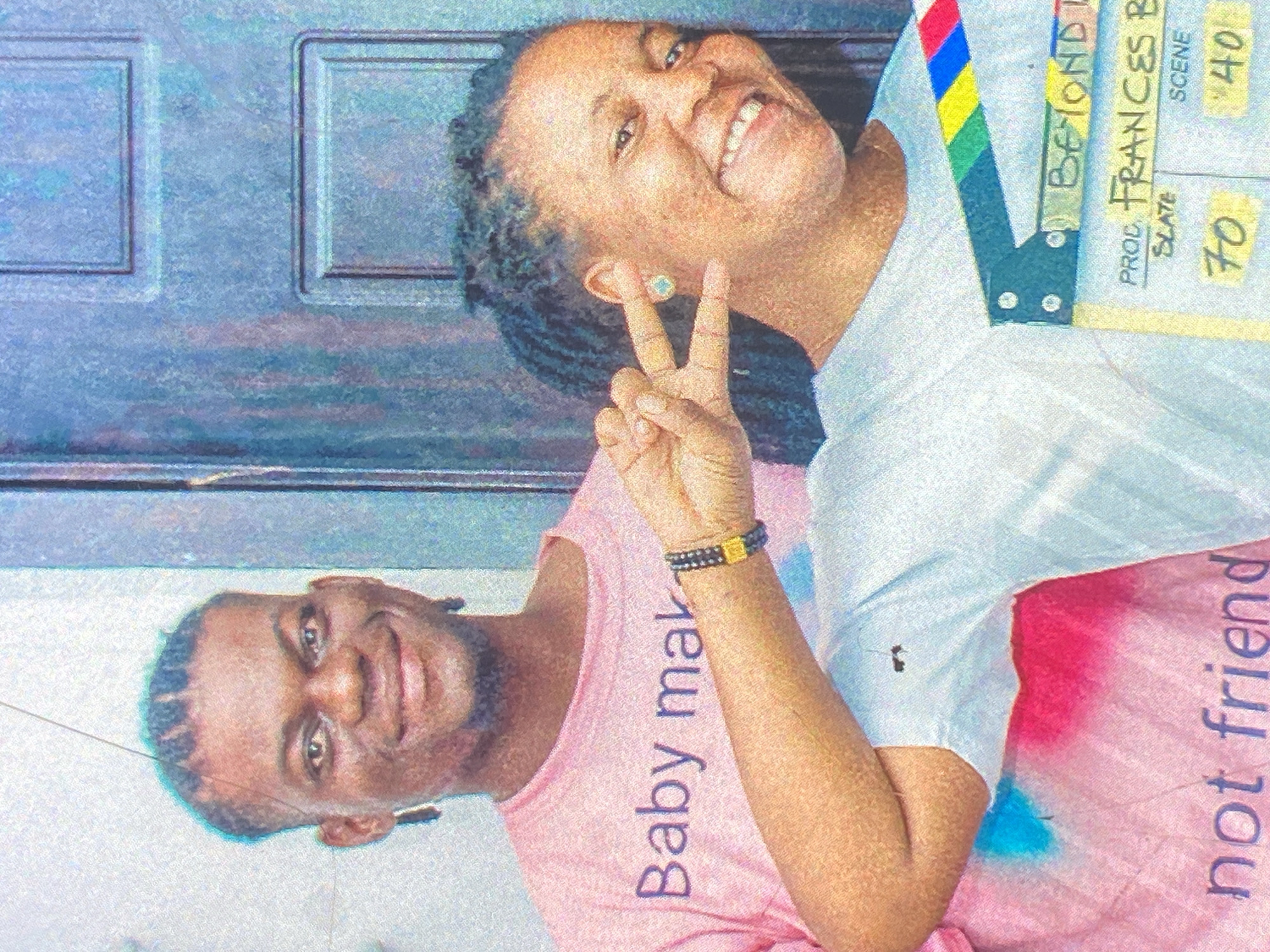 The image, rotated for proper viewing, displays two smiling individuals, a woman and a man, captured in a casual setting. The woman, positioned towards the top right, has dark, braided hair and wears a white t-shirt. She smiles broadly at the camera, making a peace sign with her right hand, and has a light blue stud earring.
Partially visible behind her and to her right is a film clapperboard featuring colorful red, yellow, green, and blue stripes. The clapperboard displays text including "BEYOND" (partially visible), "PROD FRANCES B", "SLATE 70", and "SCENE '40'".
The man, in the lower left of the frame, also smiles widely, wearing a pink t-shirt and a dark, beaded bracelet with a gold accent on his left wrist. He has short, dark, textured hair. On his pink t-shirt, the vertically oriented text "Baby mak" is visible, likely part of a phrase like "Baby maker". On the woman's white t-shirt, near the bottom right, the vertically oriented text "not friend" is visible, and her shirt is adorned with pink and blue color splashes.
The background features a light blue paneled surface, resembling a door or wall, suggesting an indoor or sheltered outdoor location. Both individuals exude happiness and a friendly demeanor. The image has a slightly grainy or artistic filter applied, giving it a textured appearance.