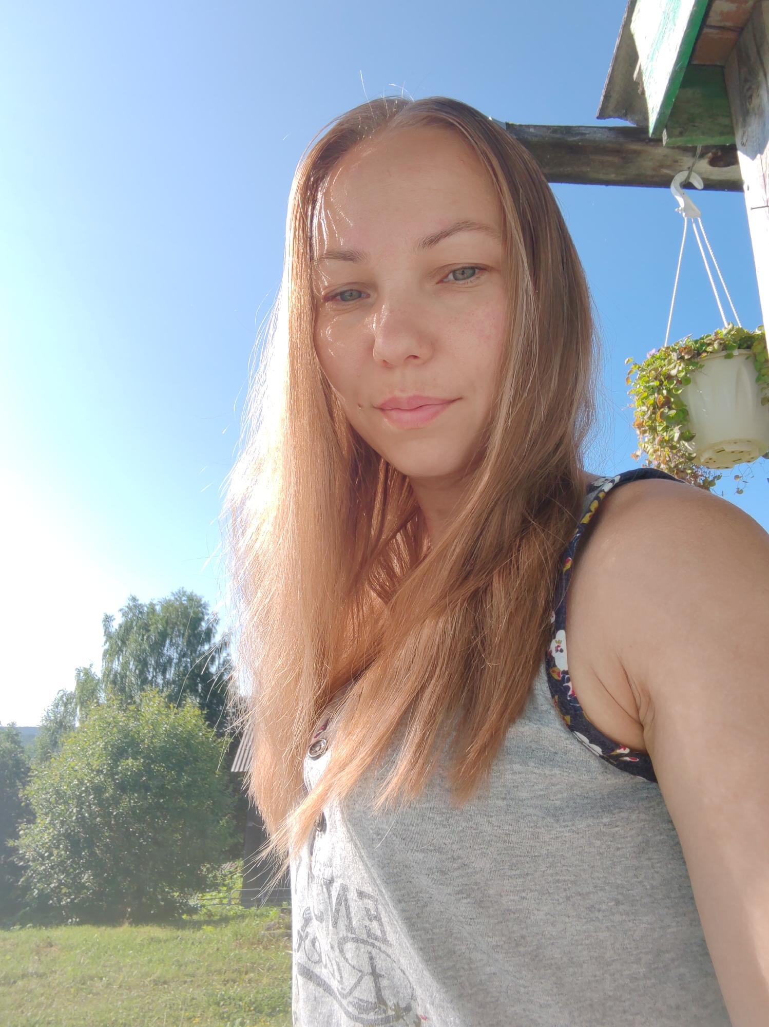 A medium shot selfie taken outdoors under a clear blue sky shows a young Caucasian woman with light brown hair and light blue eyes. She is wearing a grey t-shirt with a floral trim. The woman is smiling slightly and looking directly at the camera. In the background, there is a wooden structure with a hanging plant. To the left, there are trees and a grassy field, suggesting a rural or suburban setting, possibly in the city of Chukhlem, Russia, given the context. The lighting indicates it is daytime, likely sunny.