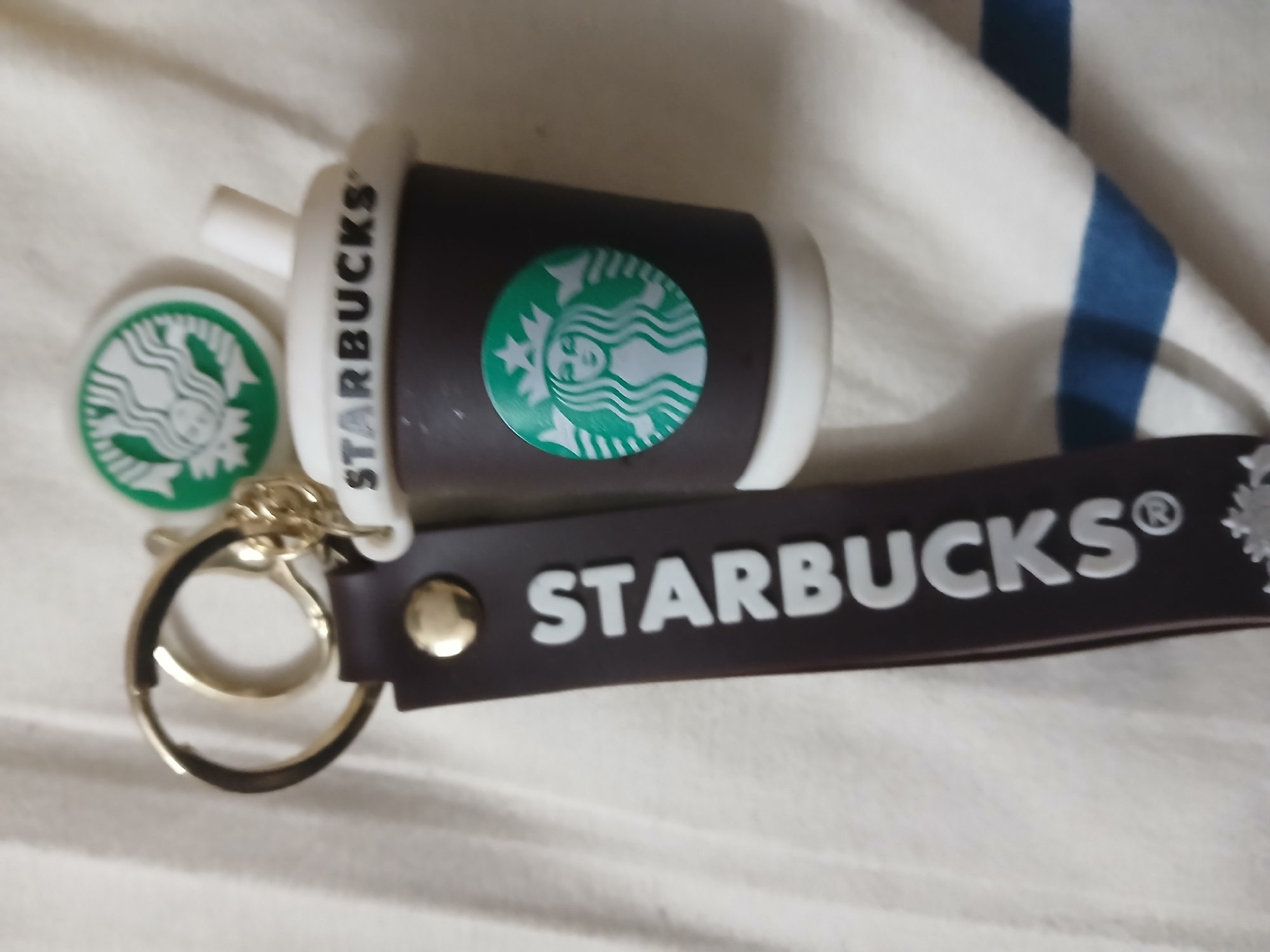 The image shows a collection of Starbucks-branded items. The main objects are a small, brown and white Starbucks cup with the Starbucks logo, a round Starbucks logo charm, and a dark brown Starbucks keychain with "STARBUCKS" printed in white. All items are placed on a white surface, possibly a bedsheet, with a blue and white striped object partially visible in the background. The environment does not reveal any information about the location in Bengaluru, India.