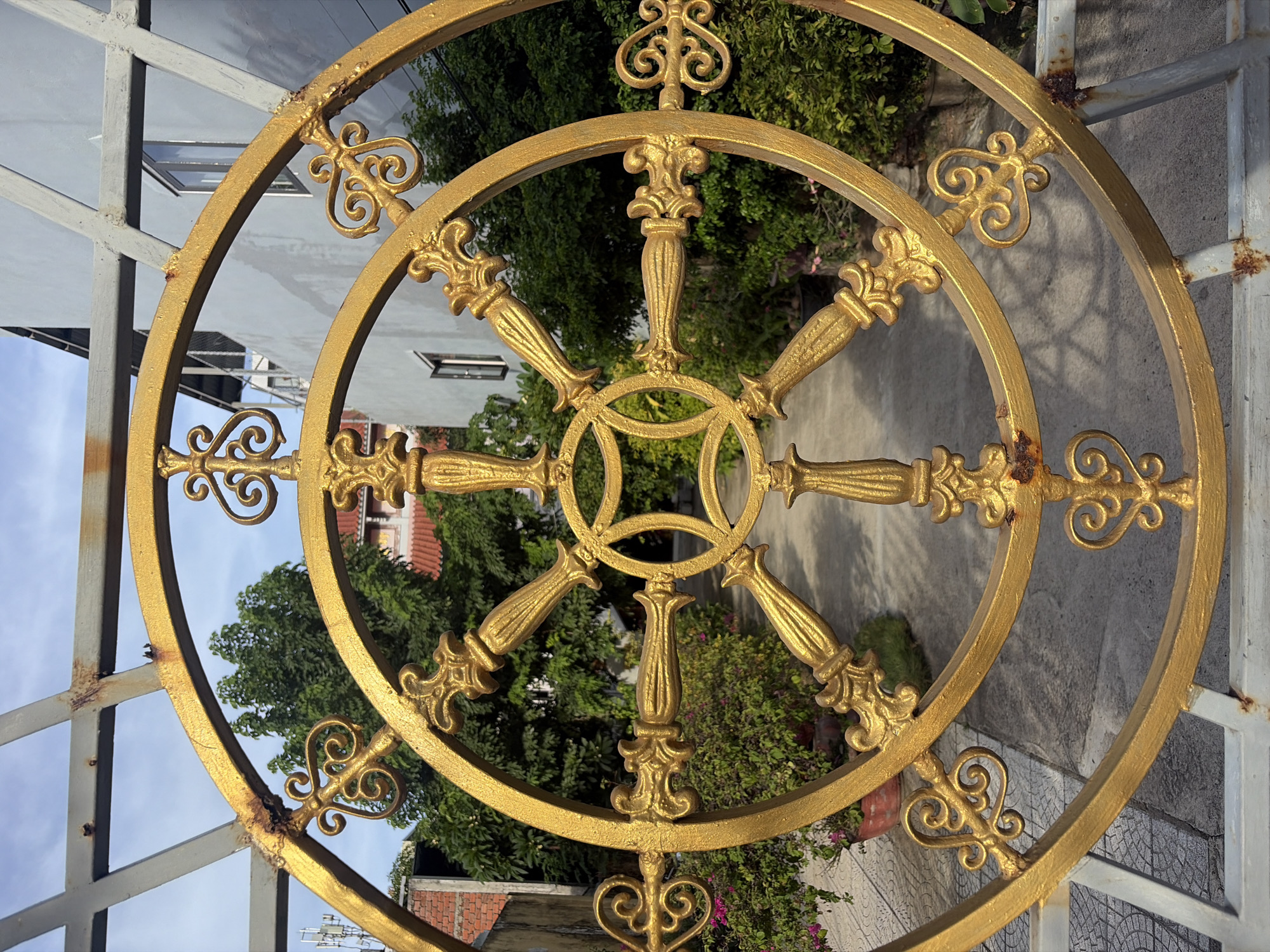 This is a close-up, eye-level shot of an ornate golden gate, possibly a Dharma wheel symbol, in Đà Nẵng, Vietnam. The gate features intricate scrollwork and decorative elements, with a circular motif at its center. Rust is visible on the gate's metal structure, suggesting it is weathered. Behind the gate, a pathway is visible, lined with lush green foliage and potted plants, some with pink flowers. In the background, there are buildings with light-colored facades and a few windows. The sky above is partly cloudy. The image is captured in daylight, with the sunlight casting shadows on the ground and highlighting the golden finish of the gate.