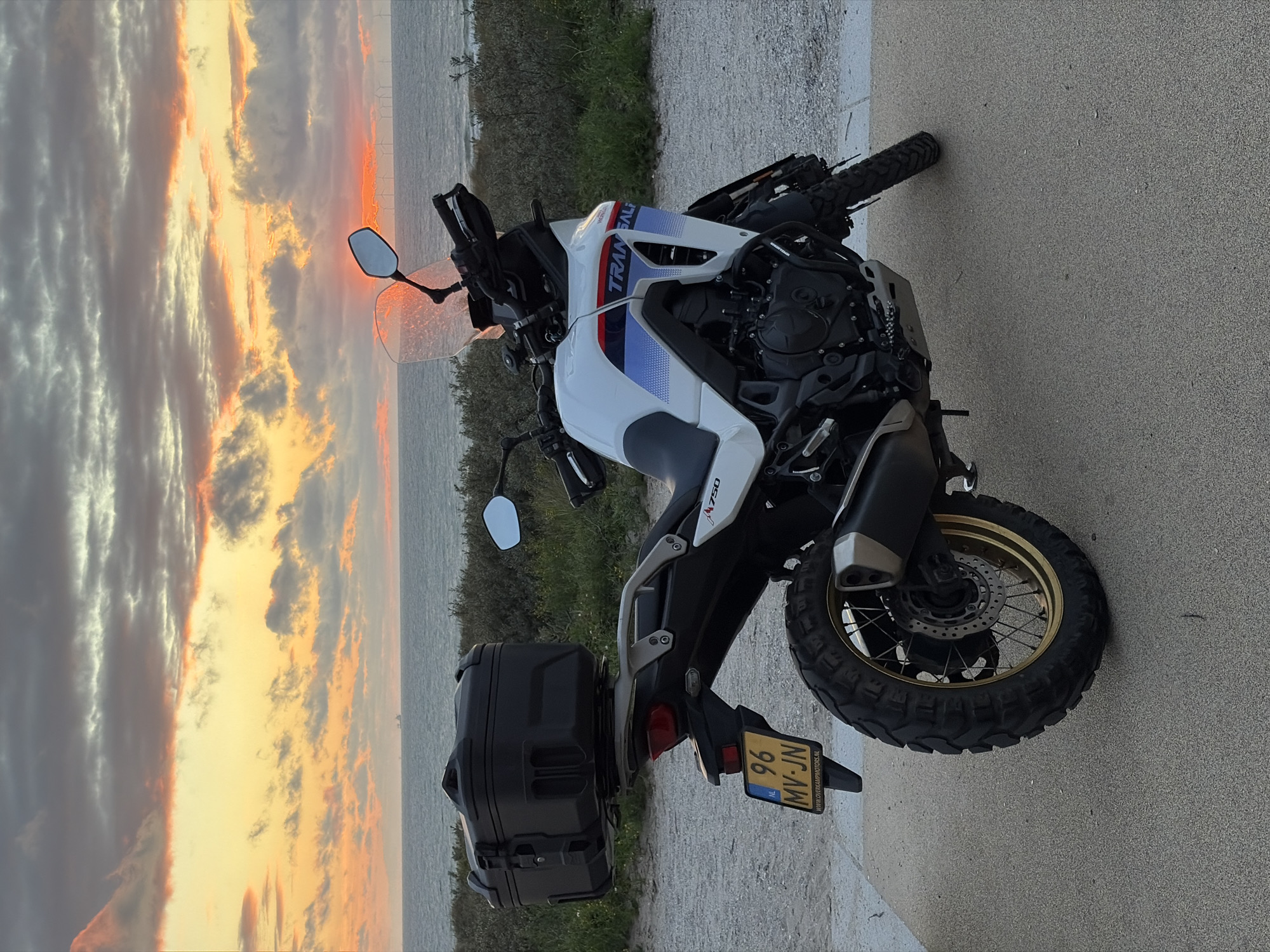 An adventure motorcycle, a white Honda Transalp 750, is parked on a paved surface next to a sandy path, with a body of water and a cloudy sunset in the background. The motorcycle is equipped with a black storage box on the rear rack and has a license plate reading "96 MV-JN" with "WWW.OVERKAMPMOTORS.NL" printed below. The surrounding environment appears to be coastal, with a hint of wind turbines visible on the horizon in the distance. The sky is filled with dramatic clouds reflecting the warm colors of the setting sun, with hues of orange, yellow, and pink. The sea is calm, stretching out towards the horizon. The ground beneath the motorcycle is a mix of sand and gravel, leading to a concrete path. The overall impression is a scenic and tranquil setting at dusk.