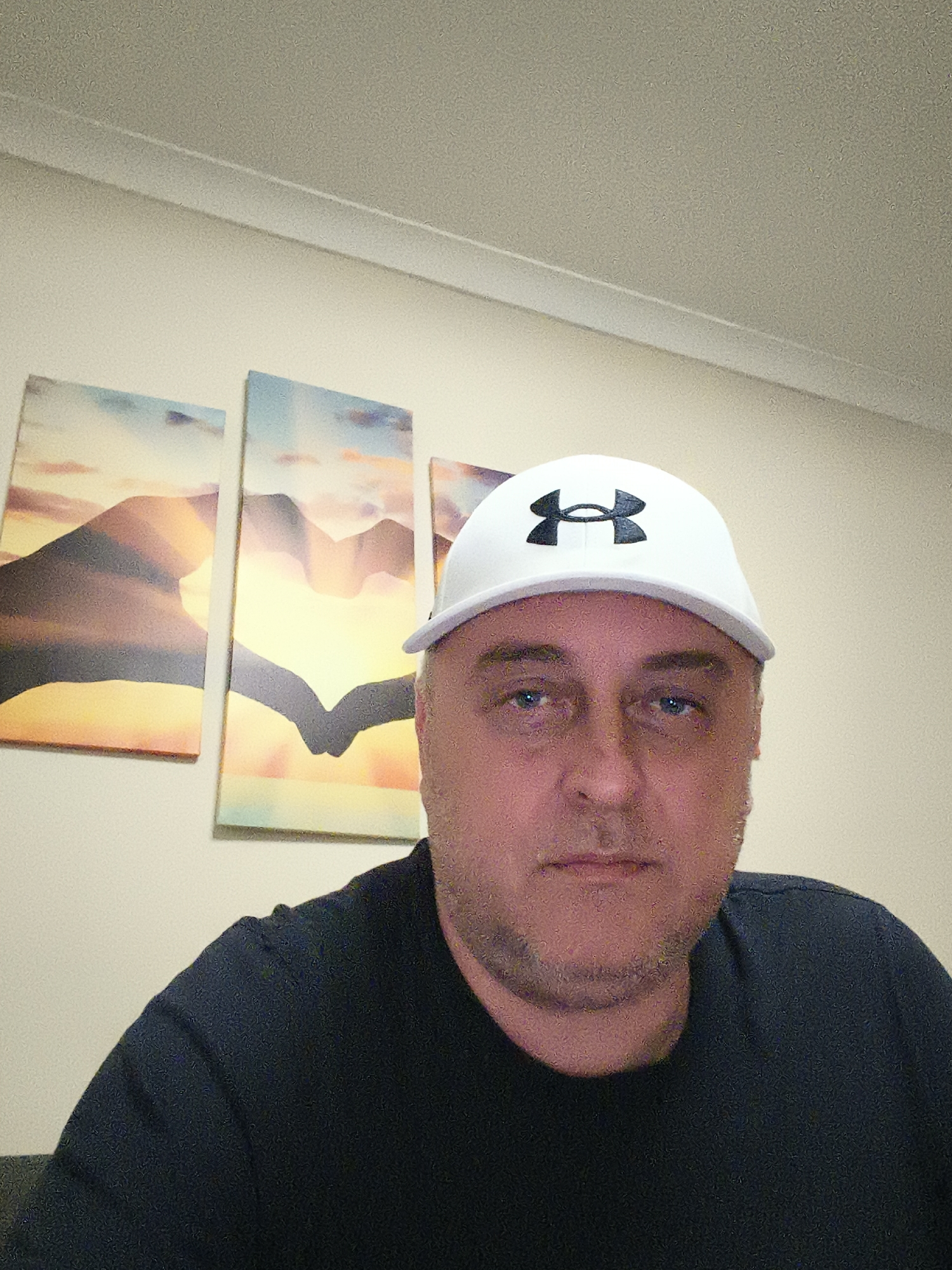 A man is featured in a head-and-shoulders portrait shot, taken indoors, likely in Huntingdonshire, United Kingdom. He is looking directly at the camera with a neutral expression. He has fair skin, blue eyes, and light stubble on his face. He is wearing a white baseball cap with the black "Under Armour" logo prominently displayed on the front. He is also wearing a plain black t-shirt.
The background consists of a light-colored, possibly off-white, wall. Along the top of the wall, where it meets the ceiling, there is a visible strip of molding. Three rectangular pieces of wall art are hung behind the man, slightly to his left. These artworks depict a stylized landscape or mountain scene with a warm color palette, suggesting a sunset or sunrise. The middle piece of art appears to show two hands coming together to form a heart shape against the bright background. The lighting in the room is even, suggesting indoor artificial light. The image has a slightly grainy quality.