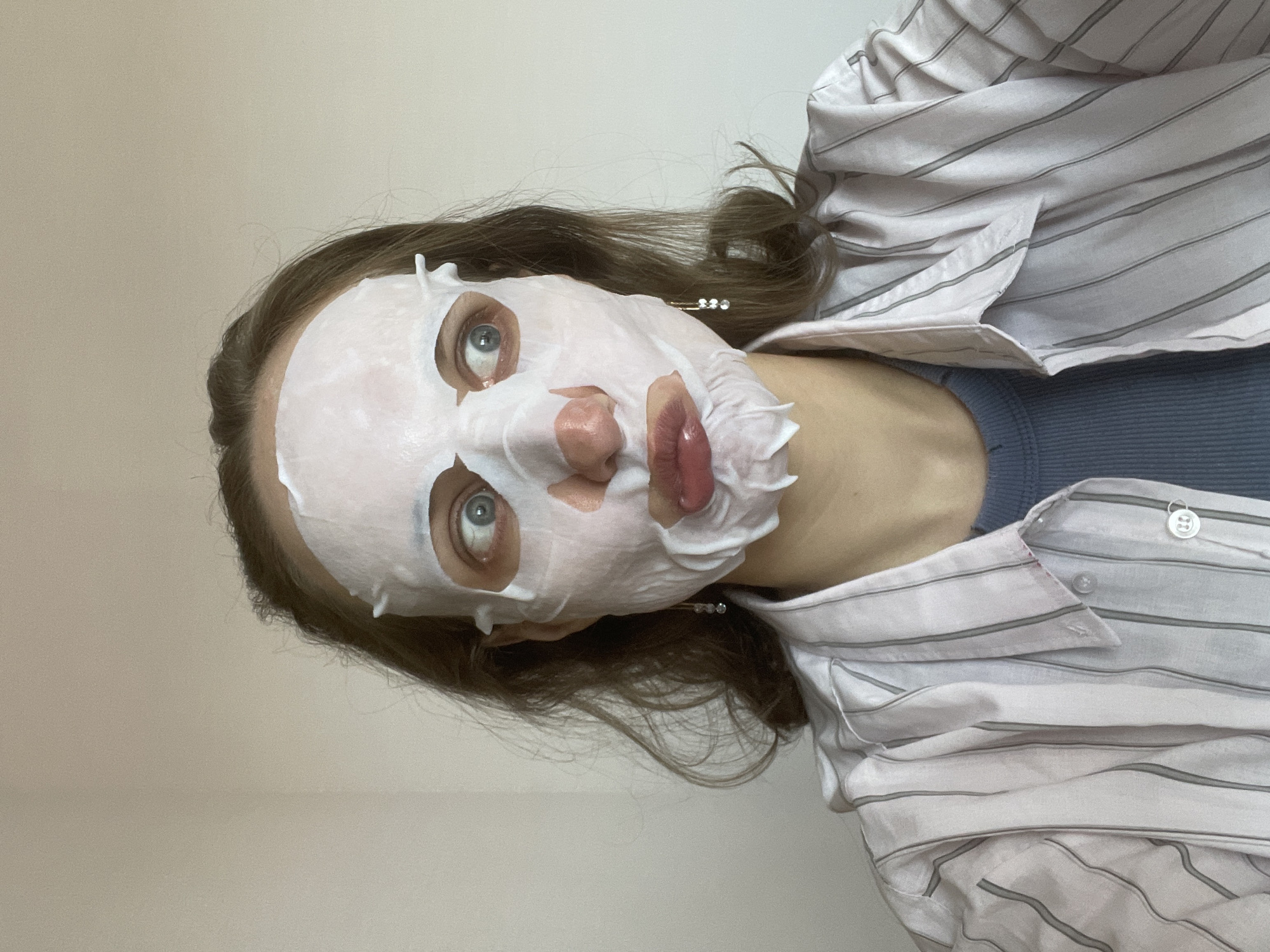 A person is shown in a close-up shot, wearing a white facial mask that covers most of their face except for the eyes, nose, and lips. The mask has a few loose sections around the nose and lips. The person has light blue eyes and brown hair. They are wearing a striped white and gray button-down shirt over a blue top. The background is a plain, light-colored wall. The location is not discernible from the image.