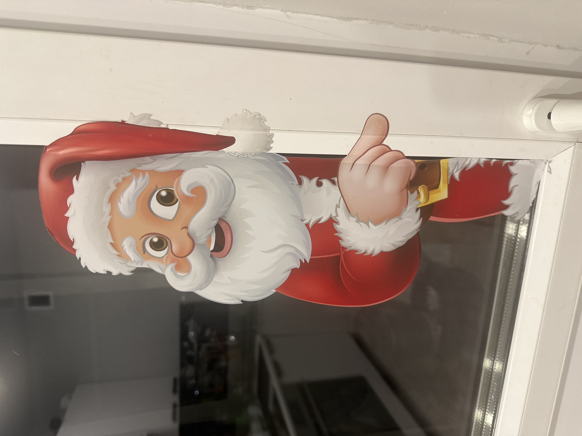 A cartoon Santa Claus sticker is adhered to a window frame. The sticker depicts Santa wearing a red hat, a white beard, and a red suit with white trim. Santa is giving a thumbs-up gesture. The window frame is white, and the reflection in the glass shows a blurry interior view. There is no information available to indicate the location is in Sochi, Russia.