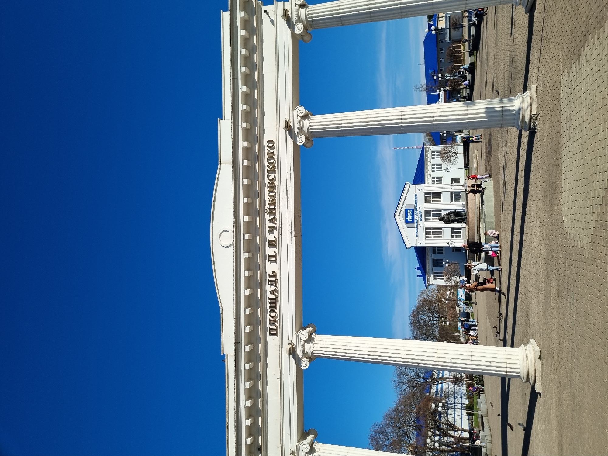 Here's a description of the image:
The image shows an entrance gate leading to a plaza in the city of Chaikovsky, Russia. The gate is made of white columns and decorative molding. The inscription on the gate reads "Площадь П. И. Чайковского", which translates to "P.I. Tchaikovsky Square". Beyond the gate is a paved plaza with people walking around. In the background, there is a white building with a blue roof and a Gazprom logo visible on the facade. There are also trees, streetlights and other buildings visible in the background. The sky is clear and blue. The image is taken on a sunny day.