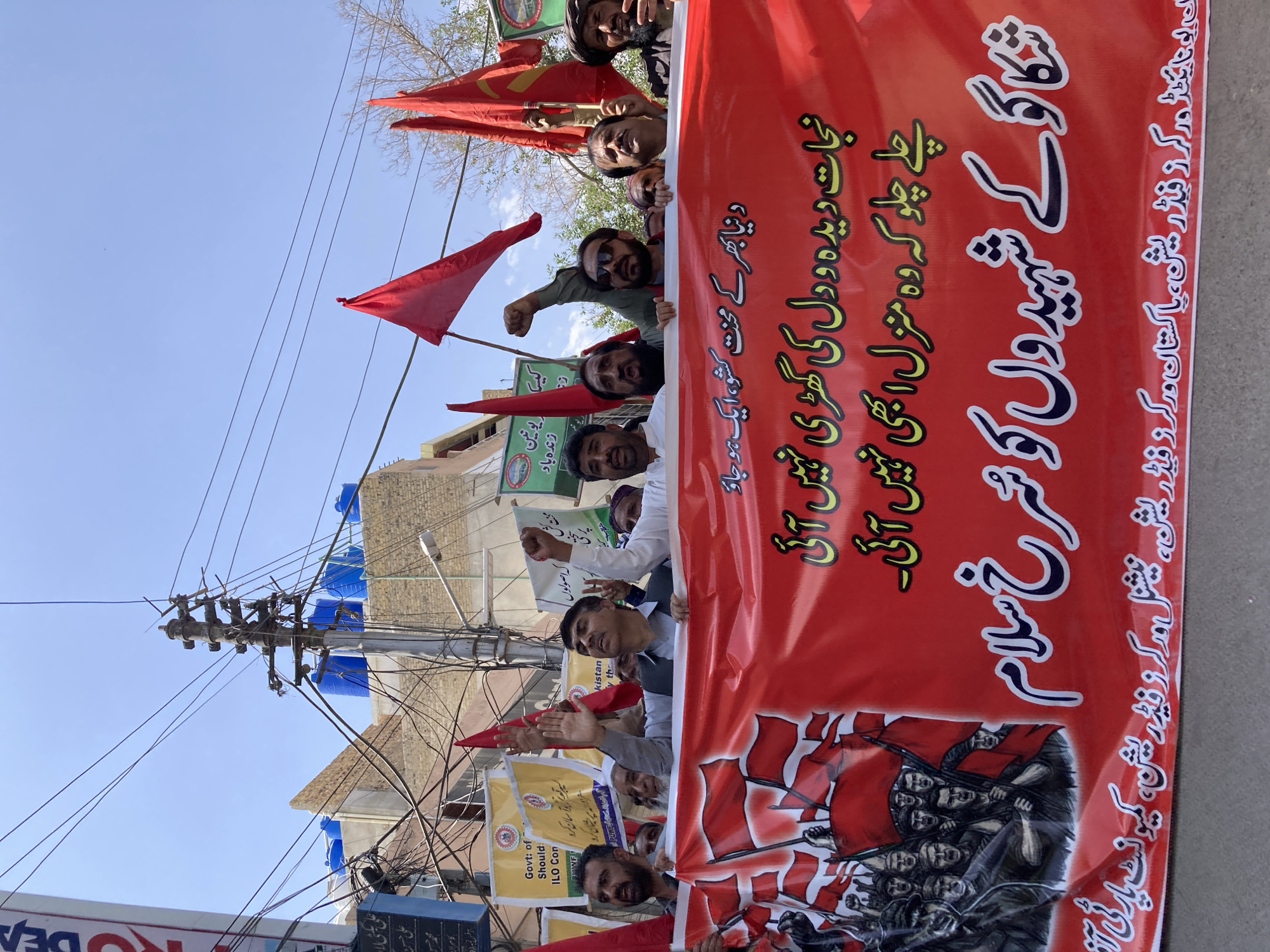 This media file captures a daylight protest or rally in an urban setting in Quetta City Tehsil, Pakistan, under a clear blue sky. A group of men, primarily young to middle-aged, are visible, holding a large red banner and several red triangular flags. Some participants are raising their fists, indicating solidarity or determination.
The central element is a prominent red banner with bold Urdu script. The main slogan at the top reads, "شکاگو کے شہیدوں کو شرخ سلام" (Red salute to the martyrs of Chicago). Below this, lines from Faiz Ahmed Faiz's poetry are written: "چلے چلو کہ وہ منزل ابھی نہیں آئی۔ نجات دیدہ و دل کی گھڑی نہیں آئی" (Keep going, for that destination has not yet arrived. The hour of salvation for the eye and heart has not yet arrived). Another visible slogan is "دنیا بھر کے محنت کشو، ایک ہو جاؤ" (Workers of the world, unite!). The banner's bottom right section credits several organizing bodies: "ان ورکرز فیڈریشن، پاکستان ورکرز فیڈریشن، نیشنل ورکرز فیڈریشن، کمیونسٹ پارٹی آ" (All Workers Federation, Pakistan Workers Federation, National Workers Federation, Communist Party of...). An illustration of figures holding red flags is at the bottom left of the banner.
Other partially visible signs include a yellow banner with English text "Govt: of Should ILO Con" (likely advocating for ILO Convention implementation) and a green banner with "کمیونسٹ پارٹی زنده باد" (Long Live the Communist Party). The background features buildings, numerous overhead power lines, and blue water tanks on rooftops, typical of a South Asian urban environment. The event appears to be a politically motivated demonstration, likely focused on labor rights and communist ideals.