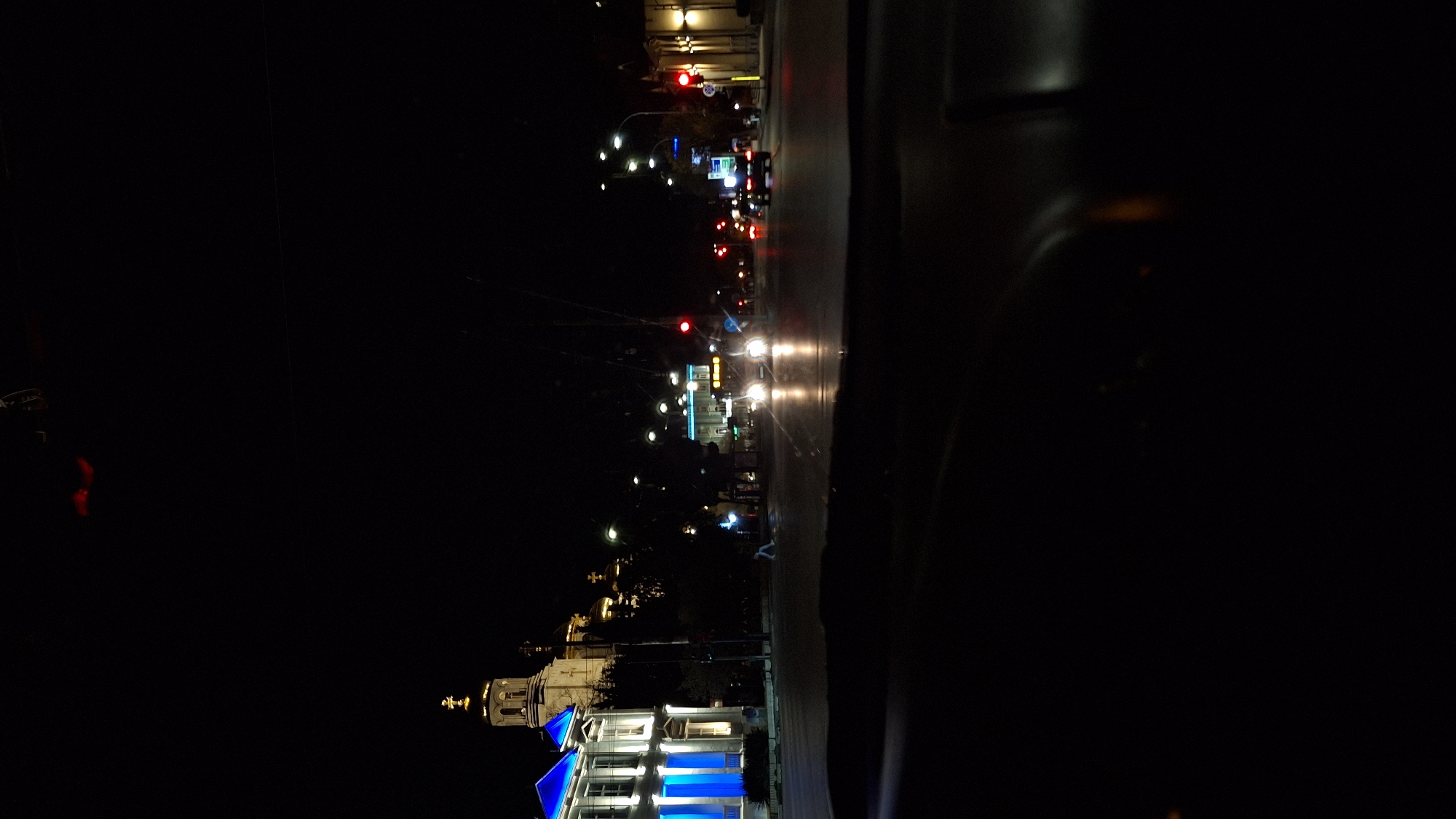 A nighttime scene shows a view through a vehicle windshield of the city of Varna, Bulgaria, from the perspective of someone driving. The street is dark, illuminated by street lights and building lights, which reflect on the wet road surface. A church or cathedral is visible on the left, brightly lit with white and blue lights. Further up the street, a row of buildings is visible. The street lights and traffic lights are also visible, with red traffic lights. Cars are moving up and down the street. The background is very dark.