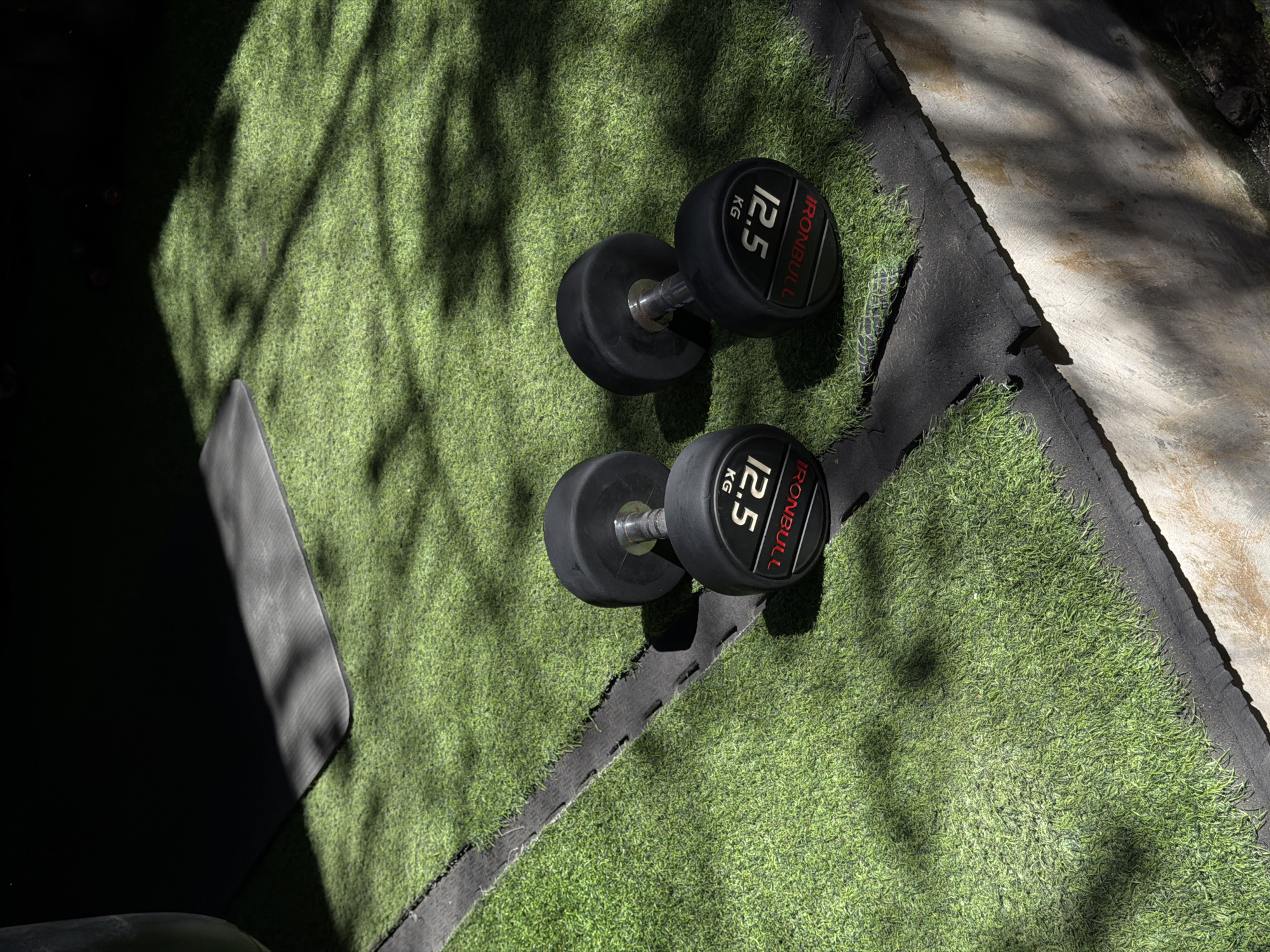 This image depicts an outdoor exercise area in North Kuta, Indonesia. The scene is dominated by artificial green turf covering the ground, interspersed with visible black rubber matting at seams and edges. Two black "IRONBULL" dumbbells, each weighing "12.5 KG" (indicated by white text on a red background), rest on their sides on the turf in the mid-ground. Both dumbbells feature chrome handles. In the upper left quadrant, a dark grey exercise mat with rounded corners is partially visible, largely obscured by a prominent dark shadow. Dappled sunlight casts distinct shadows of foliage across the turf, the mat, and a textured, light-colored concrete or stone wall running along the bottom and right edges of the frame. No individuals, animals, or active interactions are present.