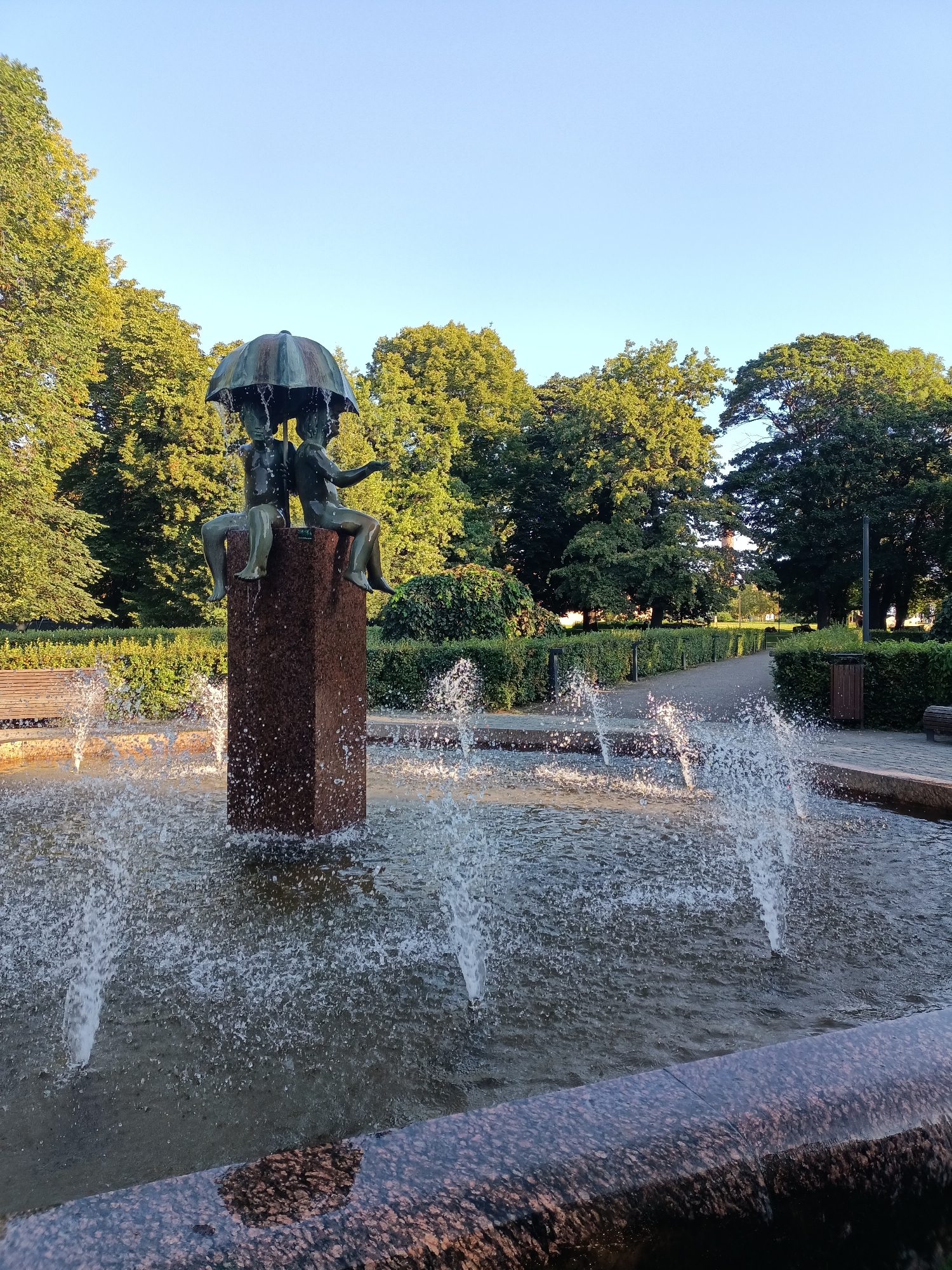 This is a medium shot, eye-level photograph of a bronze fountain sculpture in Kadriorg Park, Tallinn, Estonia. The sculpture depicts two small figures sharing an umbrella. Water streams from the umbrella and cascades into the fountain's basin. The fountain is situated amidst lush green trees and neatly trimmed bushes. In the background, a paved pathway winds through the park, with benches and trash receptacles visible along its sides. The sky is a clear, bright blue, suggesting a sunny day. The overall impression is one of a serene park setting, captured in natural light.