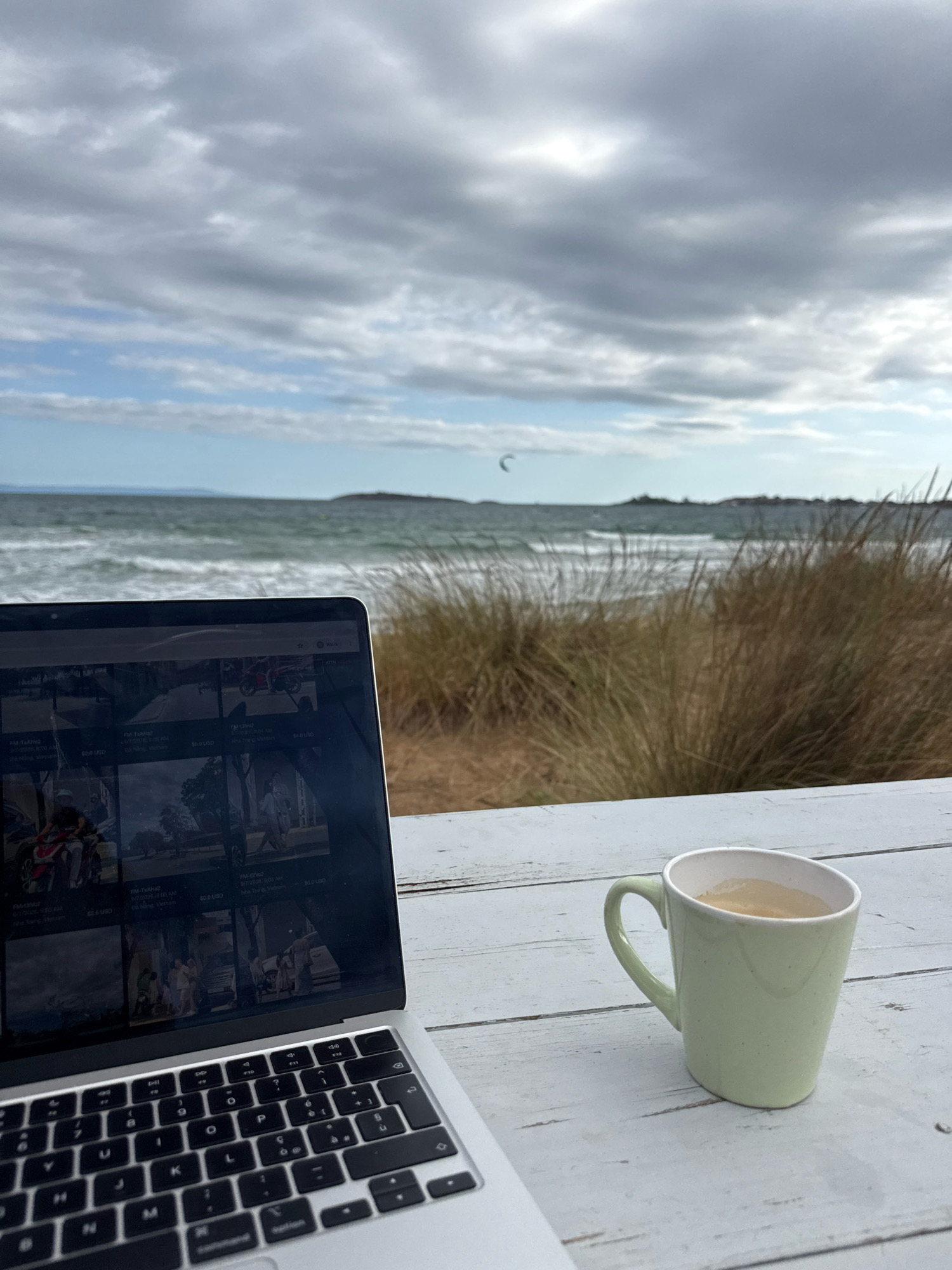 A laptop computer and a mug of coffee sit on a light-colored wooden table. The laptop screen displays a grid of images, some of which appear to be photographs of people and vehicles. The mug is light green and filled with a light-colored liquid. In the background, a beach with tall, dry grass leads to the sea. The sea is choppy with waves and in the distance, a small landmass with buildings is visible. The sky is cloudy with patches of blue. A small kite is visible in the sky over the water. The scene appears to be a peaceful outdoor setting, possibly a cafe or a rented space by the beach, in or near Sozopol, Bulgaria, given the architectural style of the distant buildings and the coastal environment.