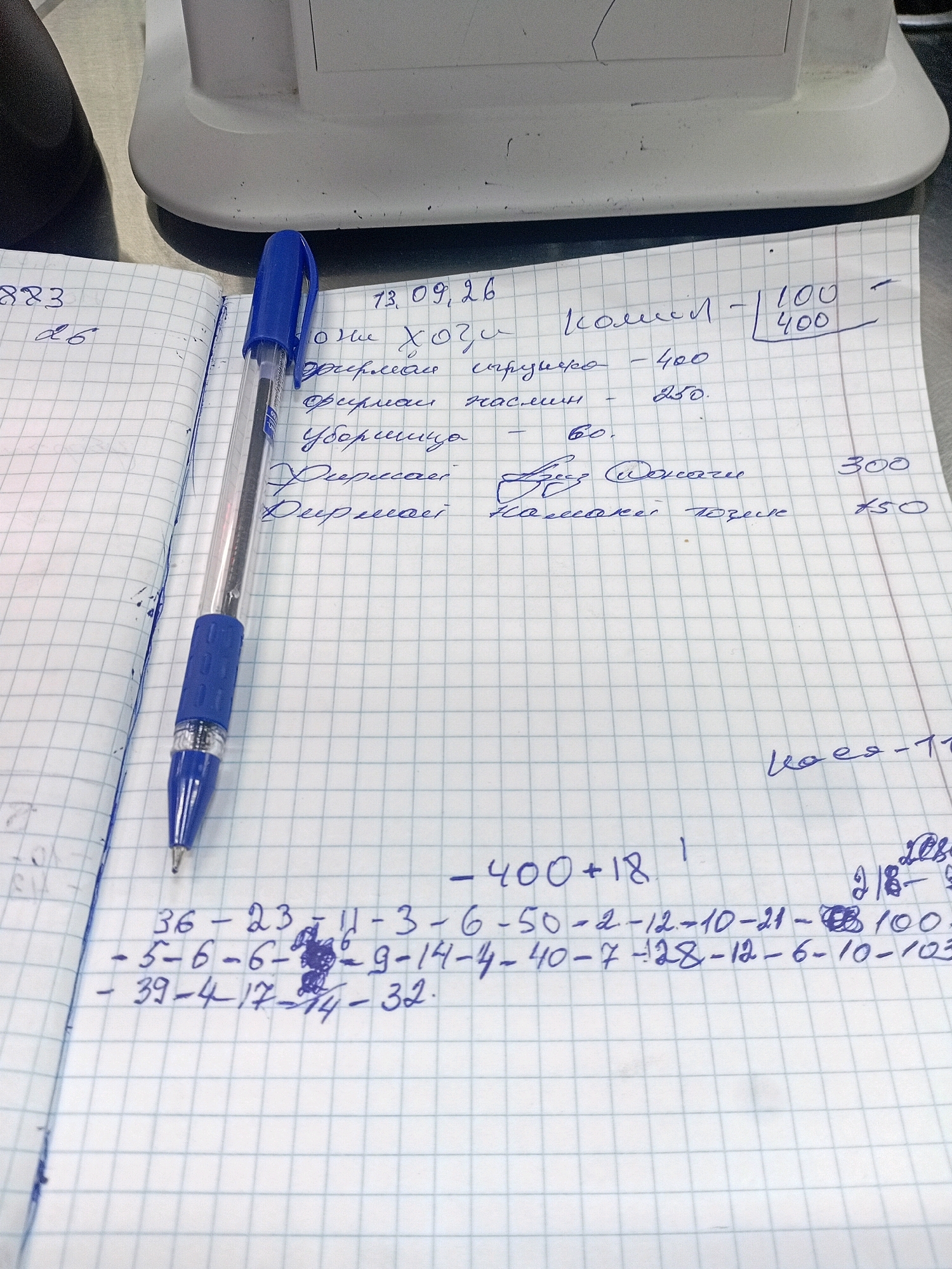 The image shows a close-up of a notebook with handwritten entries. The notebook is open, with a blue pen lying across it. The entries appear to be in Russian and include dates, numbers, and what seem to be itemized lists with corresponding monetary values. Some of the text is difficult to decipher due to handwriting and image quality. There is a white, plastic object in the background, which could be a scale or a piece of office equipment. The overall impression is of a record-keeping or financial document. The location of Dushanbe, Tajikistan is not visible in the image.