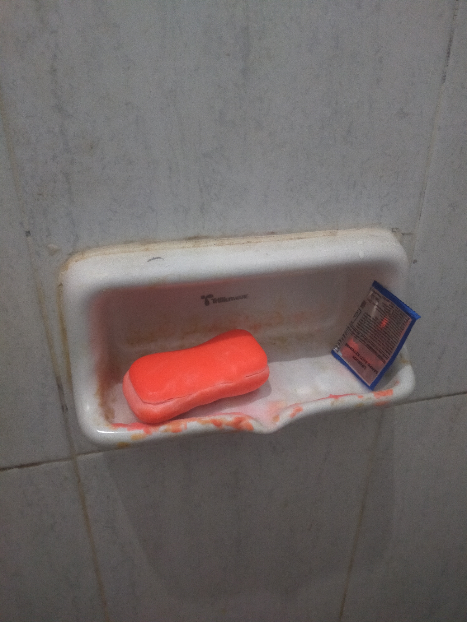 The image depicts a white ceramic soap dish mounted on a tiled wall. Inside the soap dish, there is an orange bar of soap. To the right of the soap, there is a small blue sachet with some text and an image printed on it. The soap dish and the surrounding tiles appear slightly stained. The overall environment suggests a bathroom or similar area. The location of Tegalandong, Indonesia is not visible in the image.