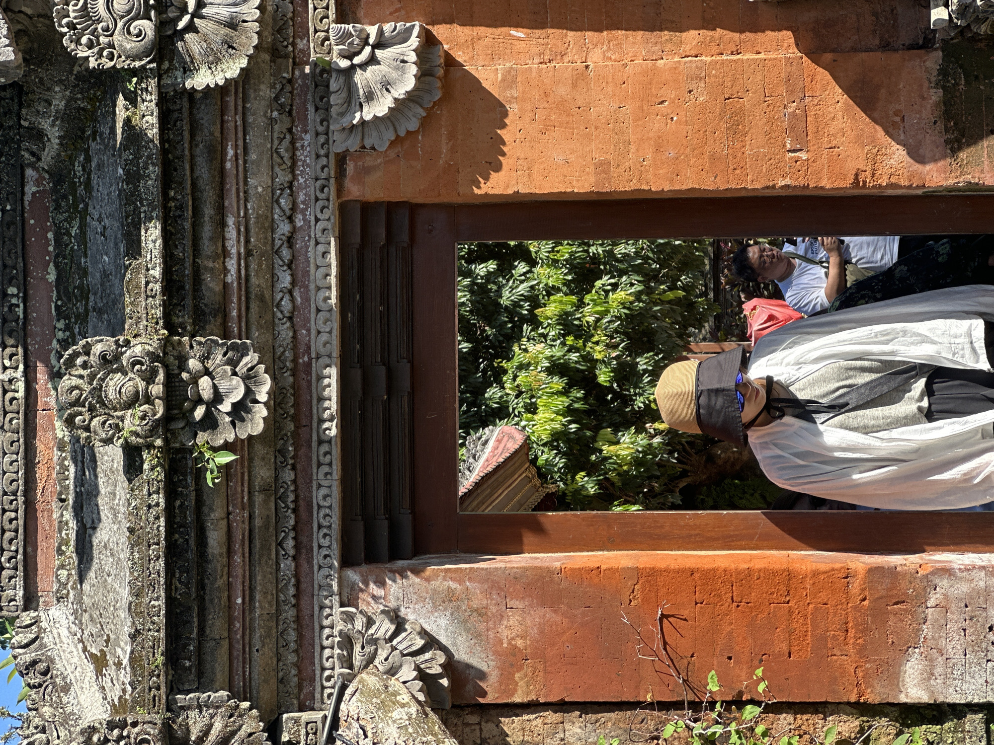In Ubud District, Indonesia, a person wearing a wide-brimmed hat and sunglasses walks through an ornate stone archway of a red brick temple. The archway is decorated with intricate carvings and is illuminated by strong sunlight, casting shadows. The person is dressed in a white long-sleeved shirt and dark pants. Behind the person, another individual wearing glasses and a white t-shirt is visible. Lush green foliage can be seen through the archway, suggesting a tropical environment. The overall scene conveys a sense of cultural exploration and a bright, sunny day.