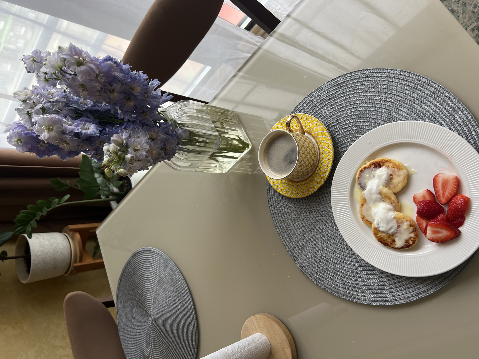 The image shows a breakfast setting on a table, presumably indoors. A vase with light purple and white flowers is positioned to the left, next to a yellow patterned cup and saucer with coffee. A white plate with small pancakes covered in white sauce and accompanied by sliced strawberries sits to the right. The plate and cup are placed on a grey placemat. The table is light beige, and a roll of paper towels rests near the bottom center. A potted plant is partially visible in the background on the left, as well as pieces of furniture.
While the location is specified as Khimki, Russia, the image provides no direct evidence to confirm this claim. The setting appears to be a private residence, possibly near a window.
