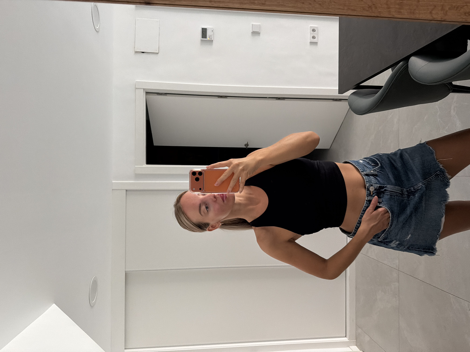 A woman with blonde hair pulled back in a ponytail, wearing a black crop top and a denim skirt, is standing indoors and taking a mirror selfie with an orange smartphone. The background shows a modern interior with white walls, sliding doors, and a doorway leading to another room. There is a dark-colored dining table and chairs visible to the right. The scene appears to be located in Finestrat, Spain.