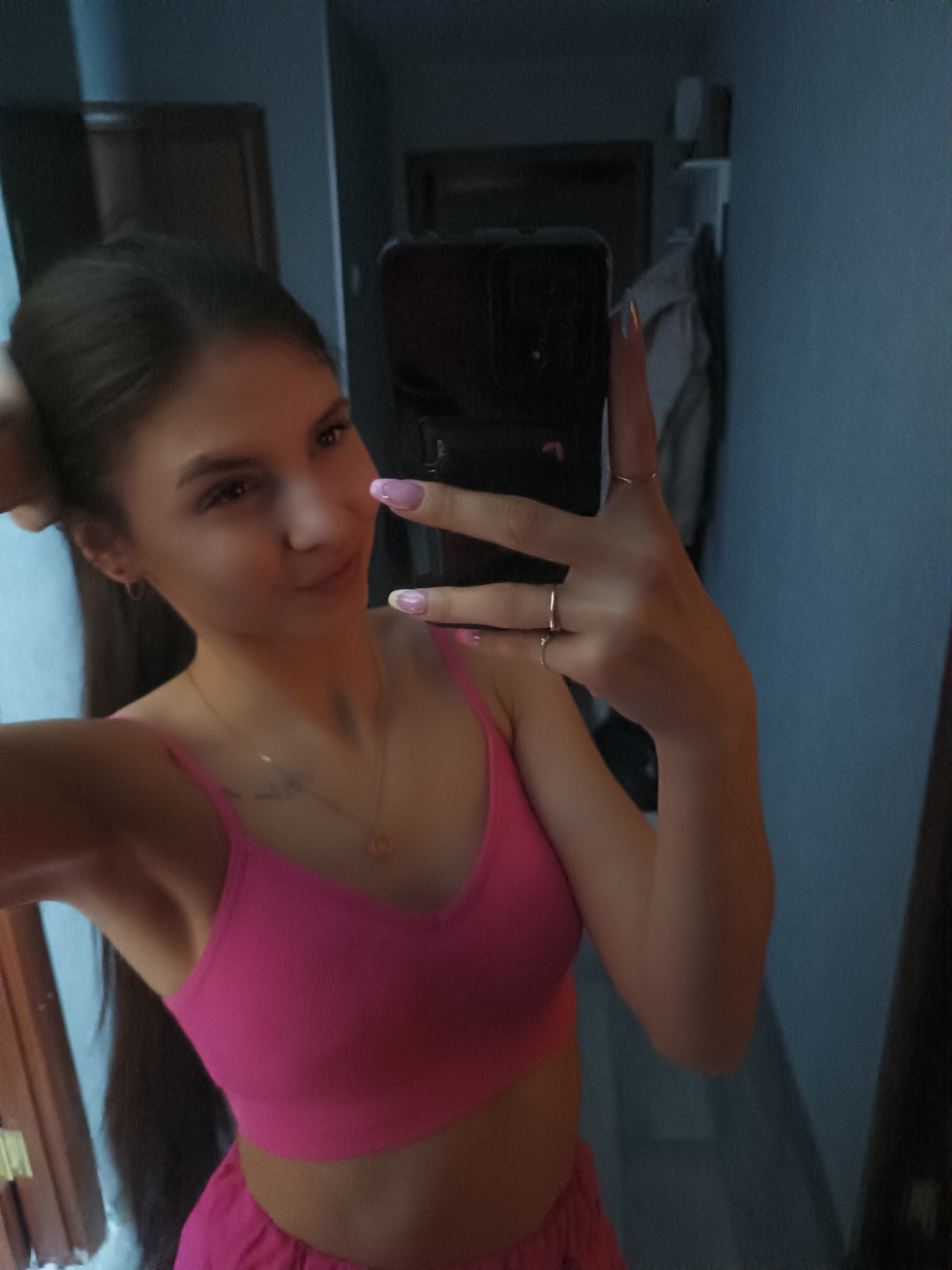 The image is a selfie of a woman indoors. She is wearing a bright pink crop top and appears to be holding her hair with one hand while taking the photo with her phone in the other. Her nails are manicured with a pink and white design, and she is wearing multiple rings on her fingers. A gold necklace is visible around her neck. Behind her, a partially visible wooden door frame is on the left, and a blurred gray wall and clothing rack with clothes hanging on it are on the right. The location seems to be a private residence, but the specific city is unknown.