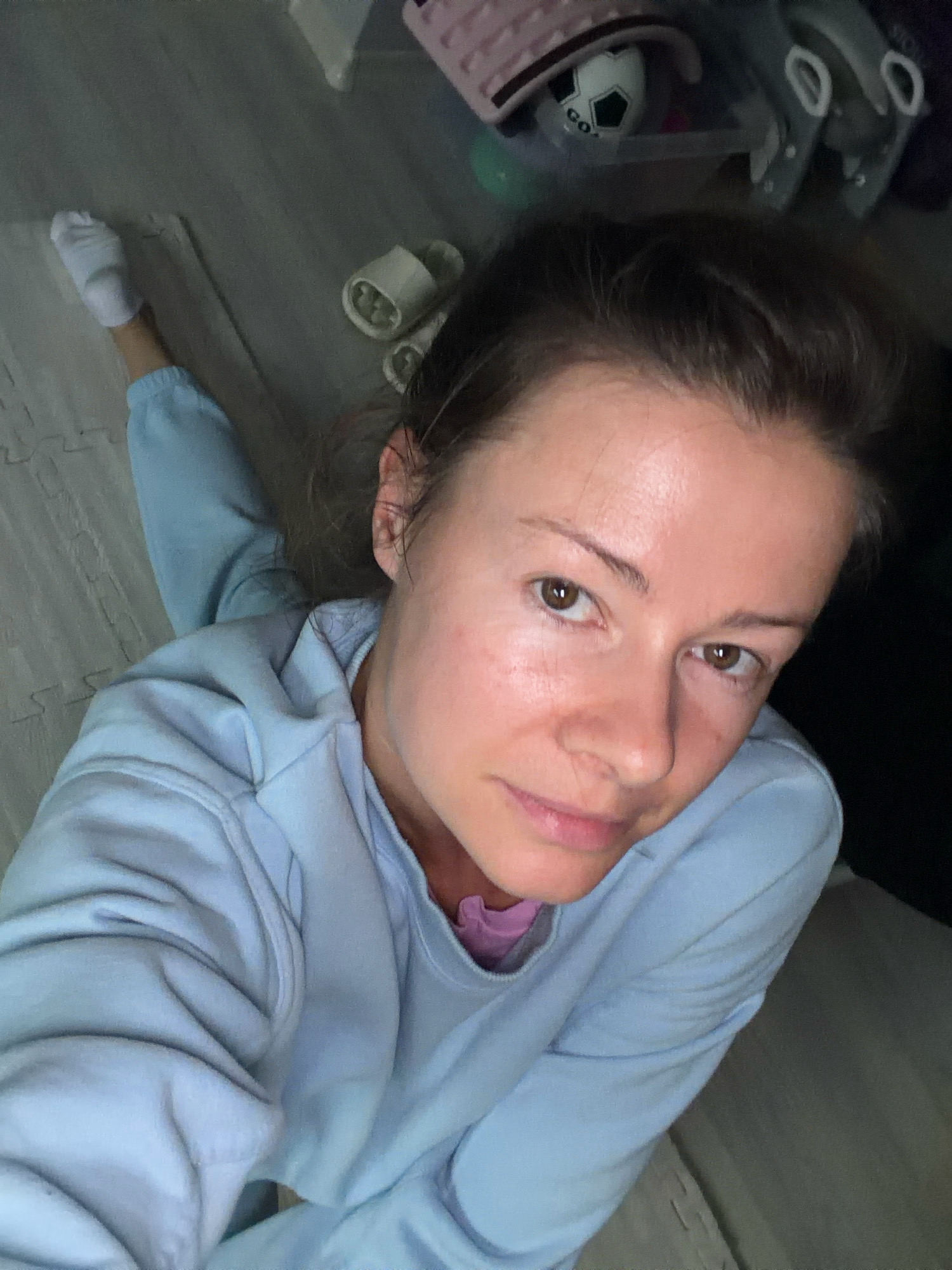 The image shows a close-up, high-angle shot of a woman in light blue loungewear, lying on a gray, padded floor, possibly a yoga mat. Her right leg is extended, wearing a white sock. In the background, there are various items scattered on the floor, including a pair of white slippers, a toy soccer ball, and some plastic containers. The lighting appears to be artificial and casts shadows, suggesting an indoor environment. The overall atmosphere is casual and relaxed. It is not possible to determine the exact location within the city of Riga, Latvia, from the provided image.
