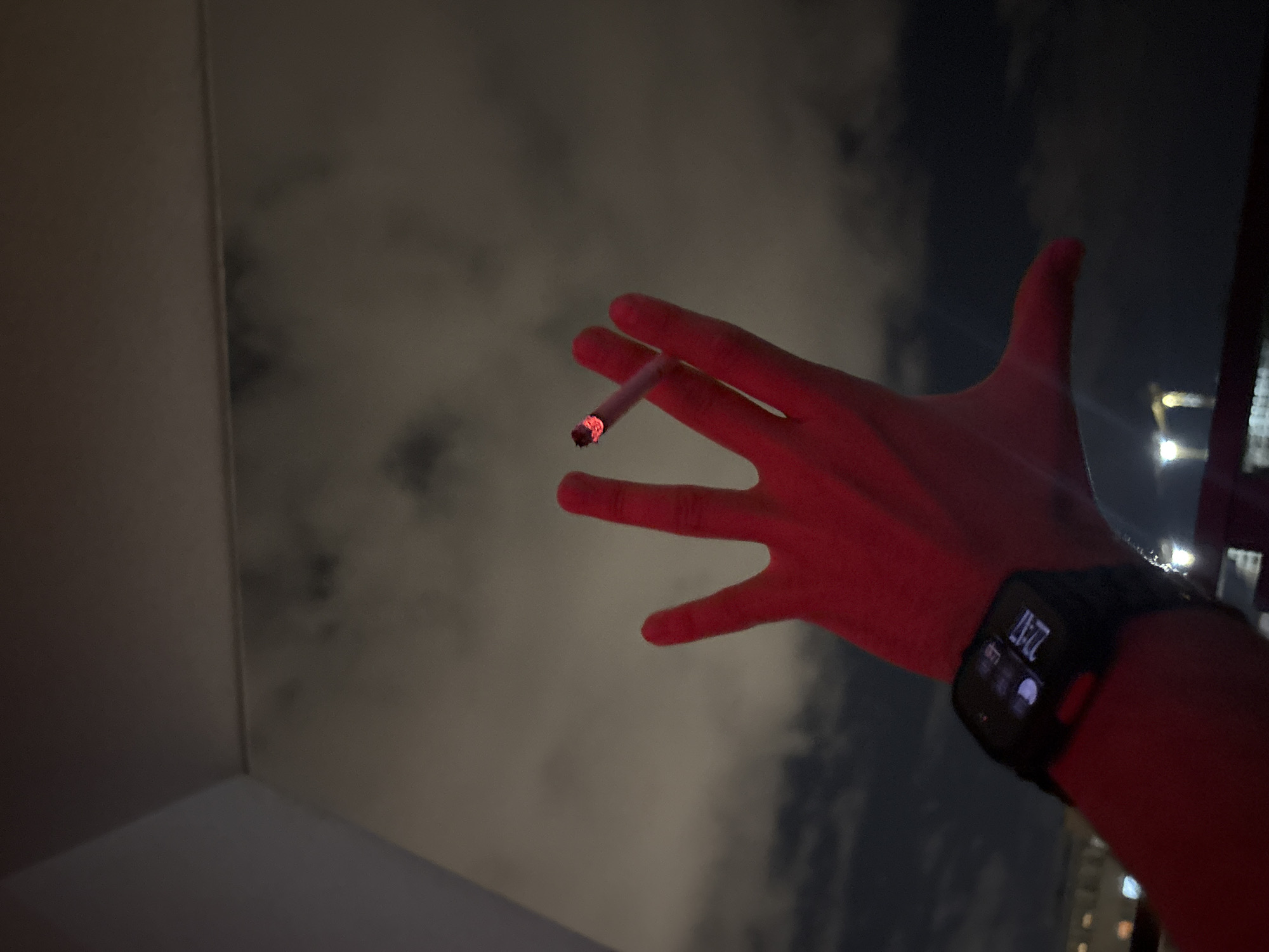 A photograph taken at night displays a human hand holding a lit cigarette. The hand is wearing a smartwatch that displays the time as 22:41. The background appears to be a cloudy night sky, with faint city lights visible, suggesting a location within an urban area. The interior corner of a building is visible in the lower left of the frame. Given the prompt's mention of Belgrade, Serbia, it is suggested that the image was taken in this city.