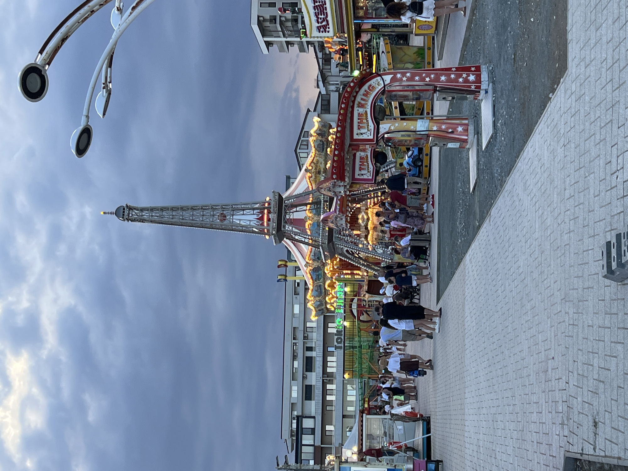 The image depicts an amusement park or fairground setting, likely located in Năvodari, Romania, given the context. In the foreground, a brick-patterned walkway extends into the scene, with a gravelly area to its right. Several amusement rides are visible, including a prominent replica of the Eiffel Tower, illuminated with lights, and a carousel with a striped canopy. A ride labeled "THE KING" with a star-spangled design is also featured. Various people are scattered throughout the park, some walking along the path, others standing near the rides. They appear to be enjoying the evening, with the sky overhead showing a cloudy, twilight atmosphere. Buildings, possibly hotels or apartment complexes, line the background. The overall impression is a lively evening at an entertainment venue.