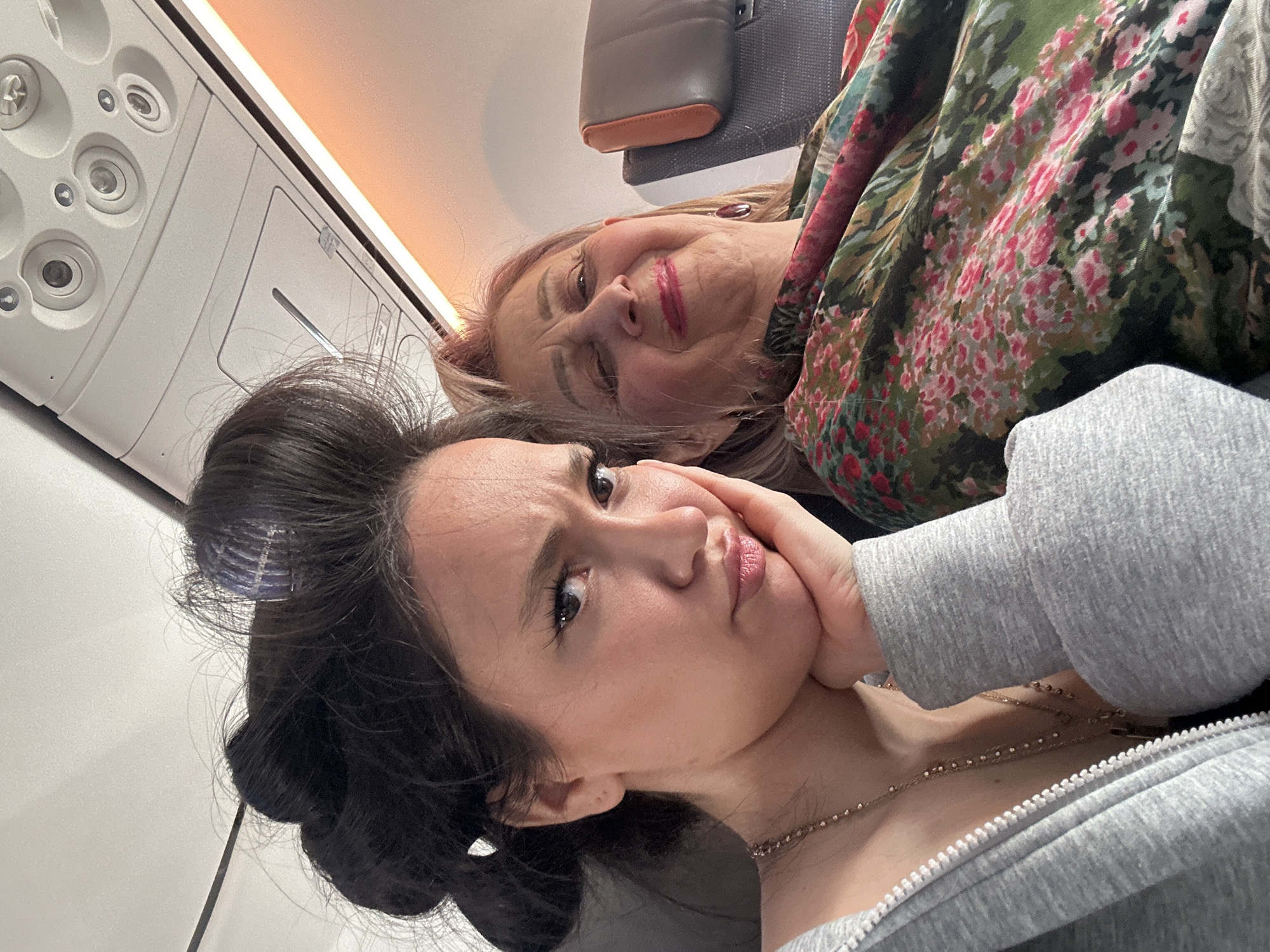 Two females are inside what appears to be an airplane. The younger female has black hair, is wearing a grey top and a gold necklace, and has her hand on her cheek. Her expression is somewhat pensive. The older female has blonde hair and is wearing a floral-patterned top. Airplane overhead panels are visible. There is no evidence indicating the city of Yerevan, Armenia, in the picture.