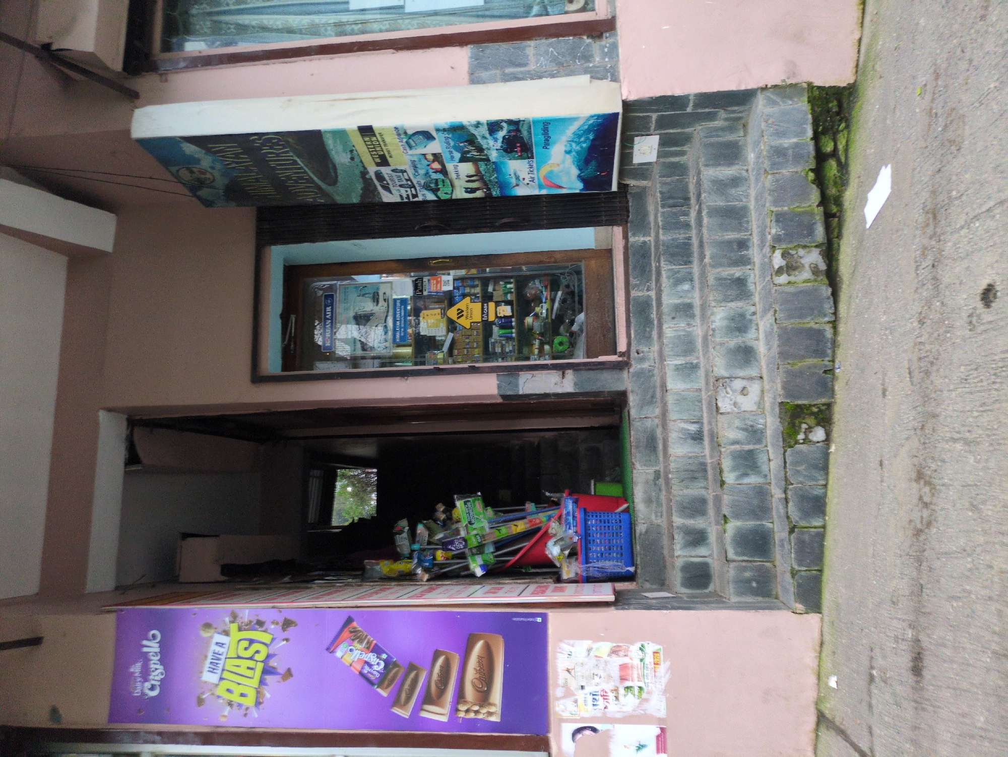 The image shows the entrance to a shop in Bhagsu, India. The shop appears to sell a variety of items, including snacks, travel-related services, and cleaning supplies. There are posters on the exterior of the shop advertising Cadbury chocolates and Himalayan adventures. Inside, shelves are stocked with various goods. A blue plastic basket filled with cleaning supplies, such as brooms and mops, is placed on the steps leading up to the shop. The steps themselves are made of stone and appear weathered. The overall scene suggests a small, local business in a bustling town.