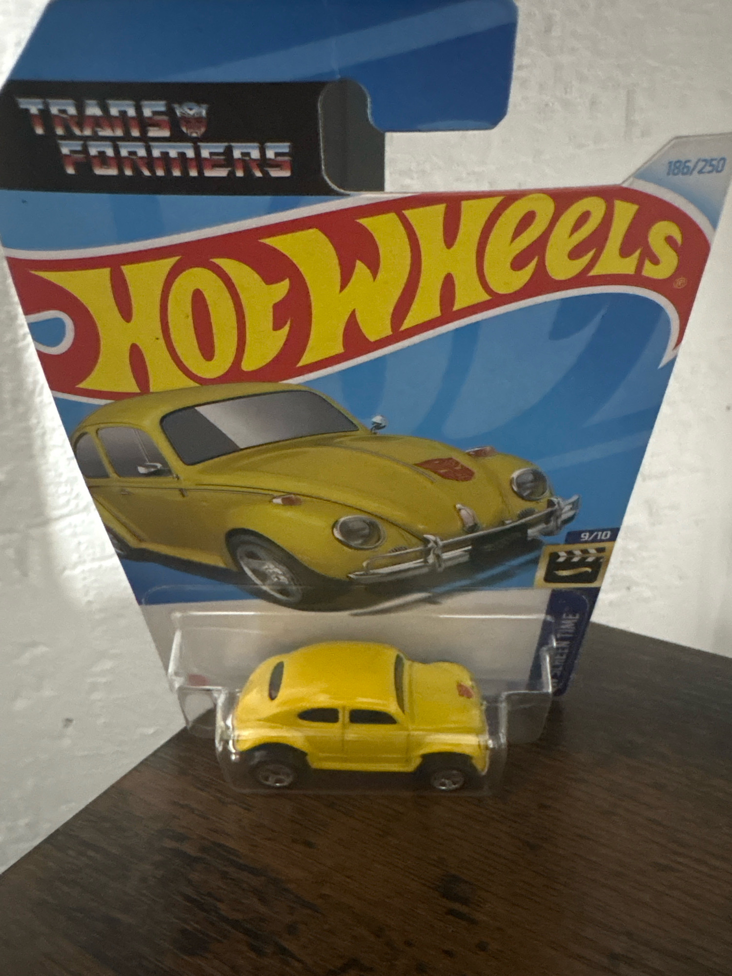 A yellow Volkswagen Beetle Hot Wheels toy car is displayed in its original packaging. The packaging features the "Transformers" logo and the "Hot Wheels" logo. A graphic of a yellow Volkswagen Beetle is on the front of the packaging, along with the text "9/10" and a clapperboard icon. The car itself is visible inside the plastic blister pack. The background is a plain white surface. The image is a medium shot, taken from a slightly elevated angle. There is no indication of location in the city of Cuernavaca, Mexico, or any people present.
