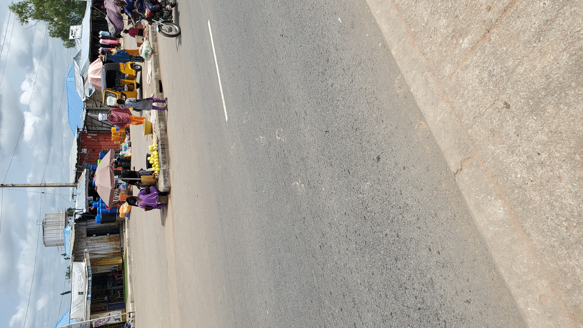 The image depicts a street scene in Jalingo, Nigeria, likely a market area. In the foreground, a paved road with a white line marking is visible, showing some light-colored smudges. The midground features a concrete sidewalk bordering the road, where several individuals are engaged in various activities. A woman in a purple outfit sits on the sidewalk, next to a pile of green fruits, possibly unripe bananas or plantains, and a yellow basin. Another person in a purple garment stands nearby. To the right, a yellow three-wheeled vehicle, commonly known as a Keke Napep, is parked. Further to the right, a person on a motorcycle, wearing a dark shirt, is visible. The background shows a row of market stalls with corrugated iron roofs, some displaying various goods like blue barrels and yellow containers. Overhead, a cloudy sky is present. The scene suggests a typical marketplace environment with vendors and shoppers.