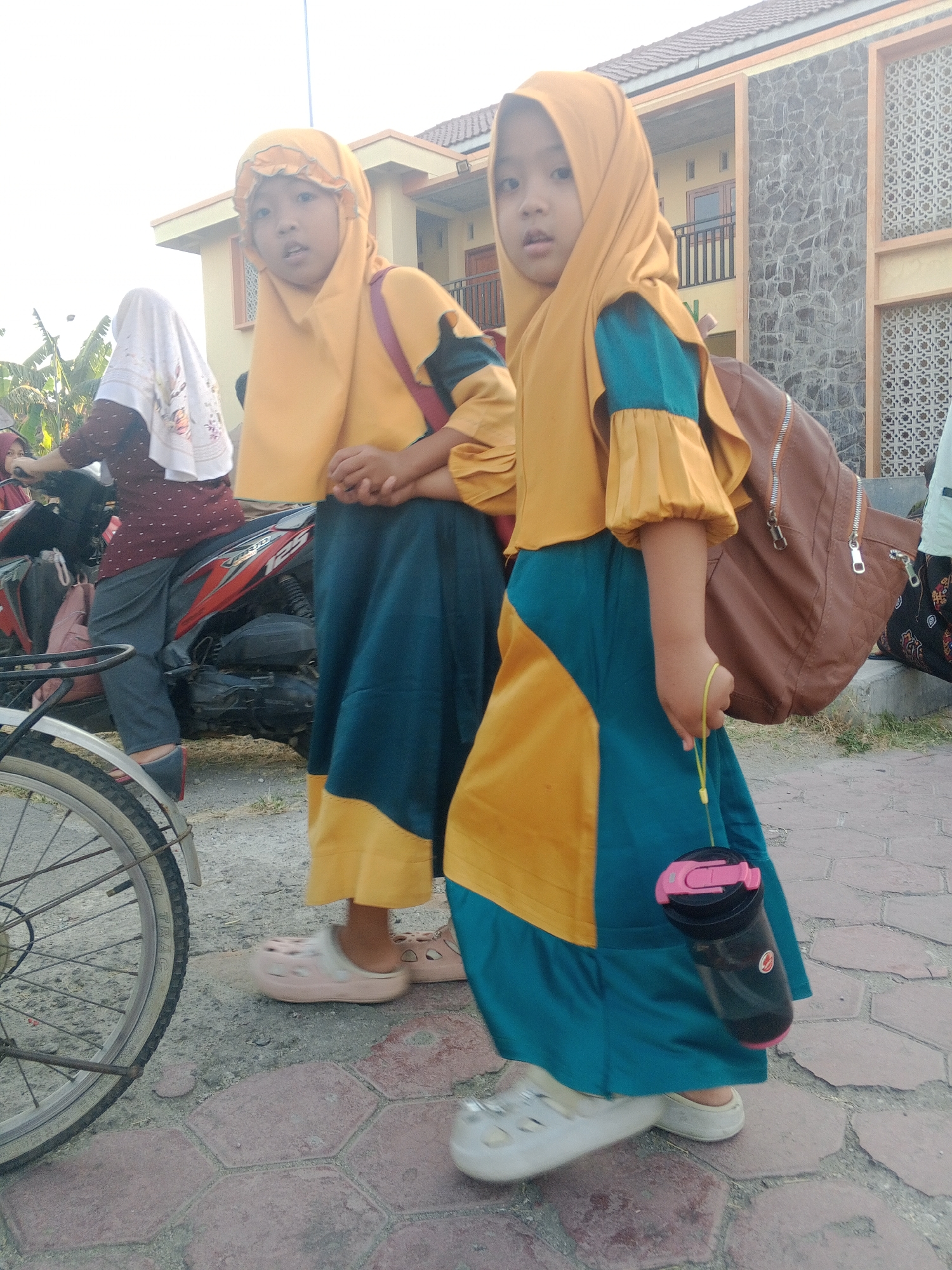 Two young girls are walking on a paved sidewalk. The girl in the foreground is wearing a yellow and turquoise dress and a yellow hijab. She is holding a black water bottle with a pink lid. The girl behind her is wearing a similar yellow and turquoise outfit with a yellow hijab. She has her hands clasped in front of her. A brown backpack is slung over the right shoulder of the girl in the foreground. In the background, a red and black motorcycle is parked. A building with a grey stone facade and light brown trim is visible behind the girls. The sky is bright and overcast. The location is identified as Lembah, Indonesia.