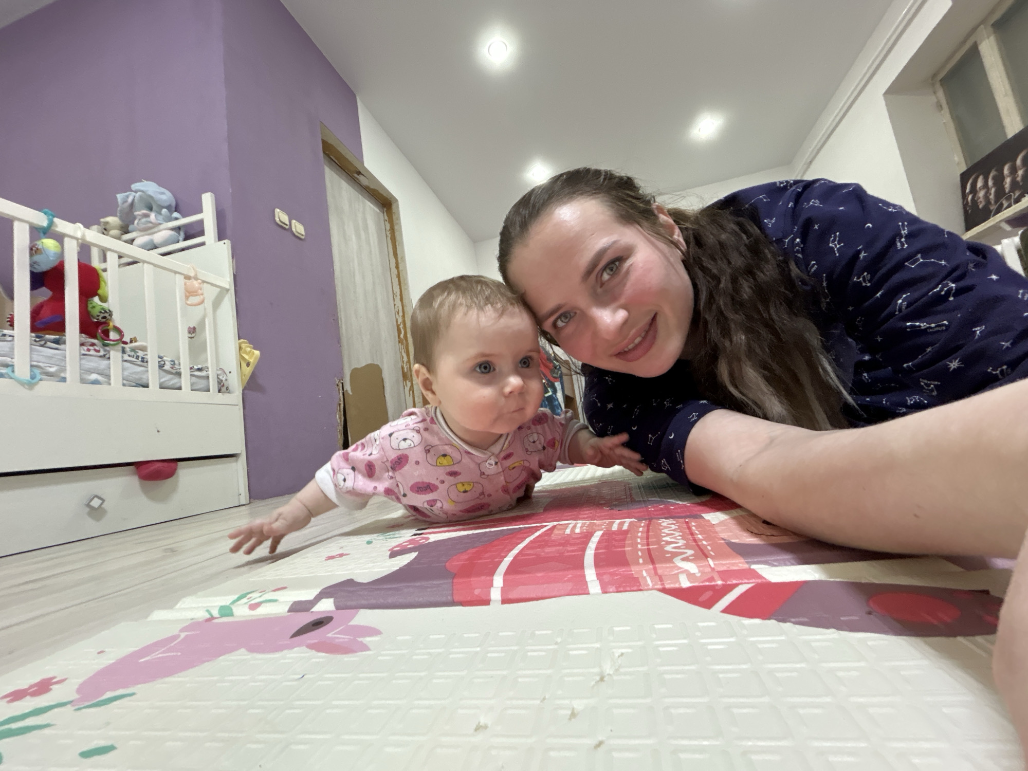 A selfie was taken from floor level in a room with purple and white walls, and light colored flooring. A baby wearing a pink patterned shirt with long sleeves is lying on its stomach on a colorful foam play mat. Next to the baby is a woman with long brown hair and wearing a navy blue shirt with white constellations on it. The woman is smiling at the camera. Behind them is a white crib with toys inside. The location is in Rusalevka, Russia.