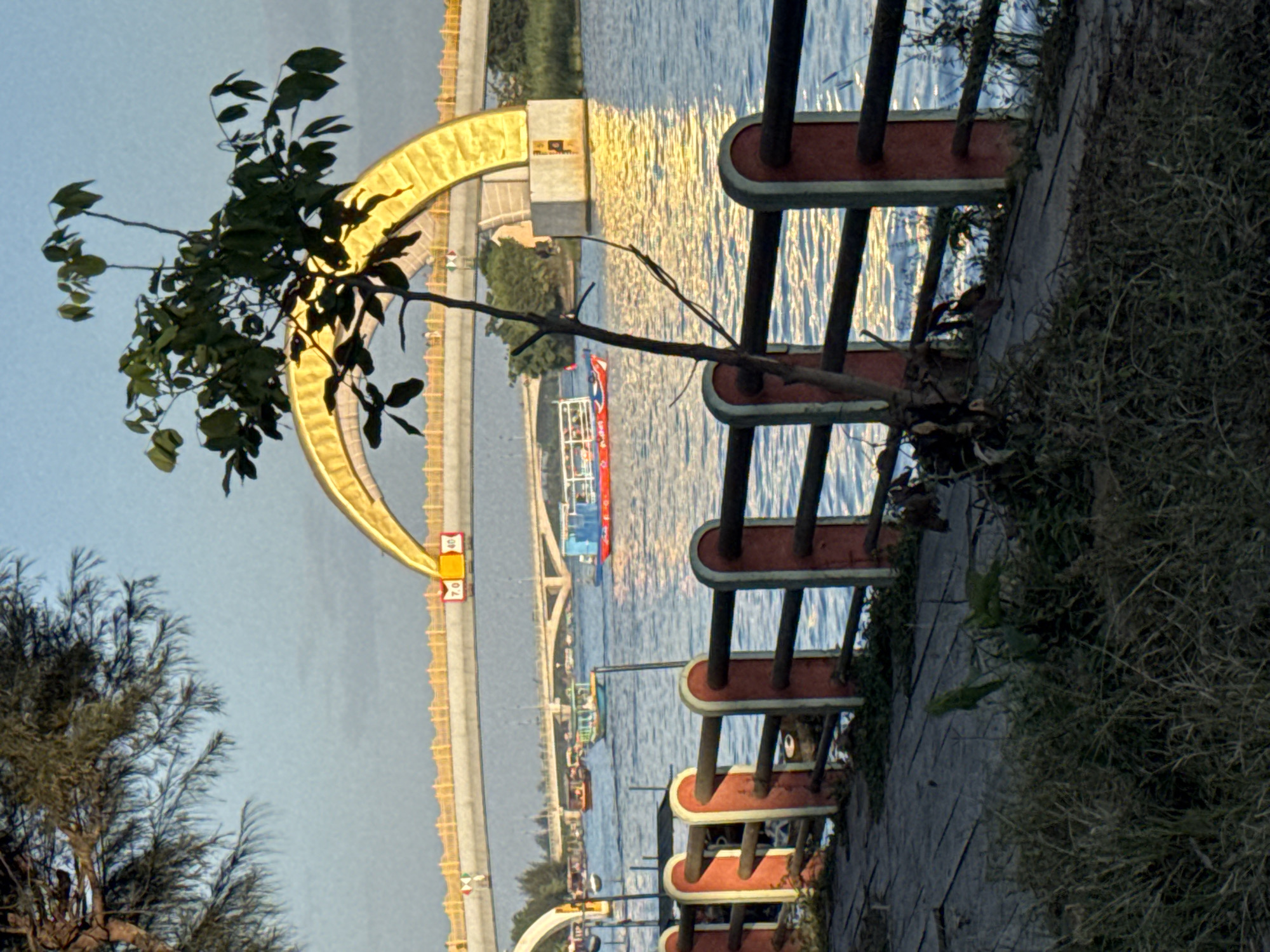 The image shows a waterfront view in Đà Nẵng, Vietnam, featuring the Trần Thị Lý Bridge with its distinctive curved yellow arch. A red and grey metal railing is in the foreground along a paved walkway with grassy overgrowth. The water shows reflective patterns of sunlight. Boats are on the water near the bridge. A tree is visible on the left side of the image.