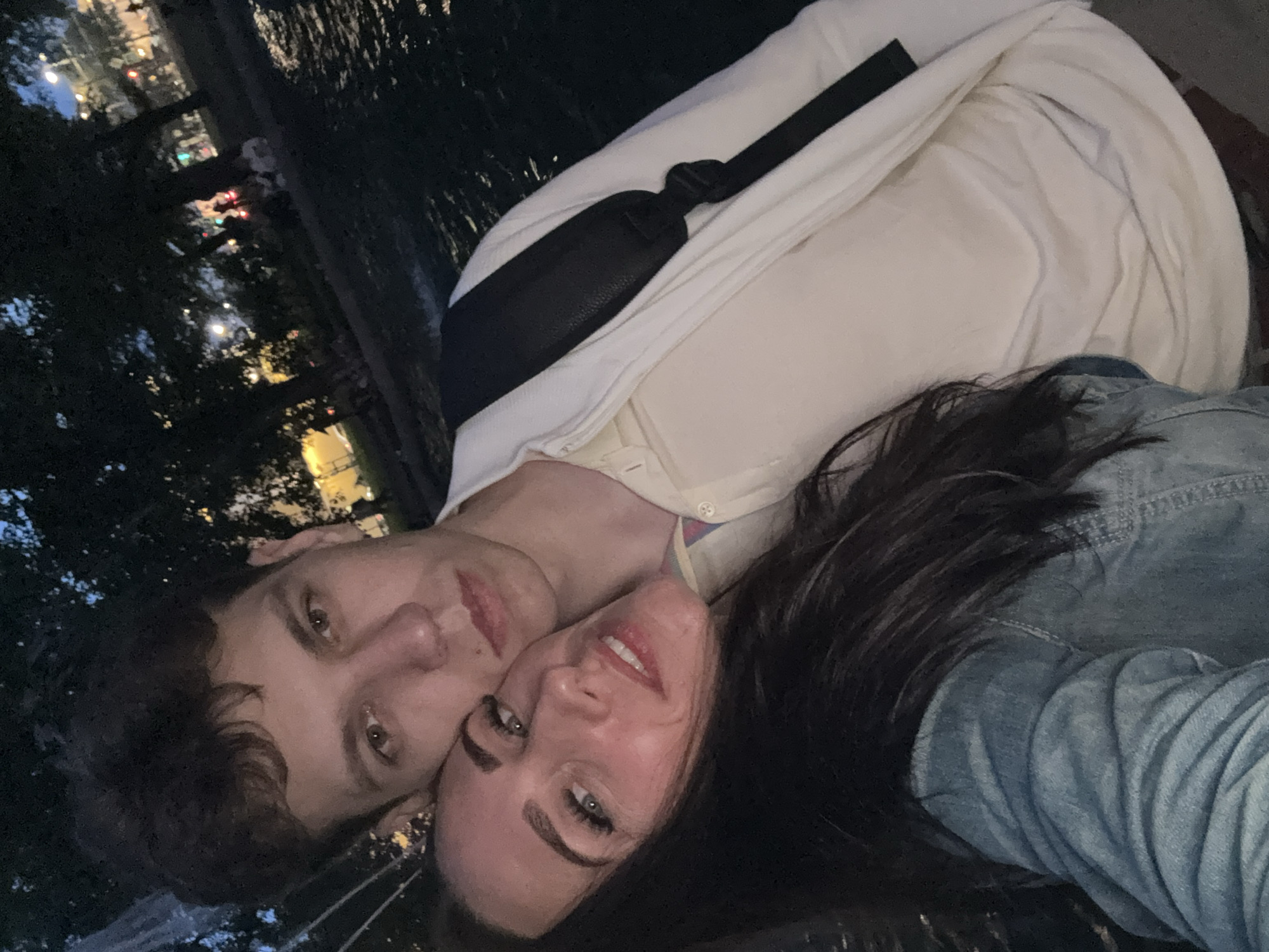 A rotated close-up portrait captures two individuals, a male and a female, likely a selfie taken at night in Saint Petersburg, Russia. They are positioned side-by-side, with their heads resting close together, looking towards the camera.
The male, on the left, has short, dark brown, curly hair and light-colored eyes. He wears a light-colored (possibly cream or off-white) collared shirt, and a black strap, likely from a bag or fanny pack, crosses his chest. His expression is neutral and calm.
The female, on the right, has long, dark hair and light eyes, with visible mascara enhancing her eyelashes. She is dressed in a light blue denim jacket or shirt. Her lips are slightly parted in a soft, relaxed expression.
The background is dark and out of focus, indicating a nighttime setting outdoors. Blurred artificial lights are visible in the distance, along with indistinct dark foliage, suggesting an urban environment, potentially near a body of water common in Saint Petersburg. The scene conveys a quiet, intimate moment between the two individuals. No visible text is present in the image.