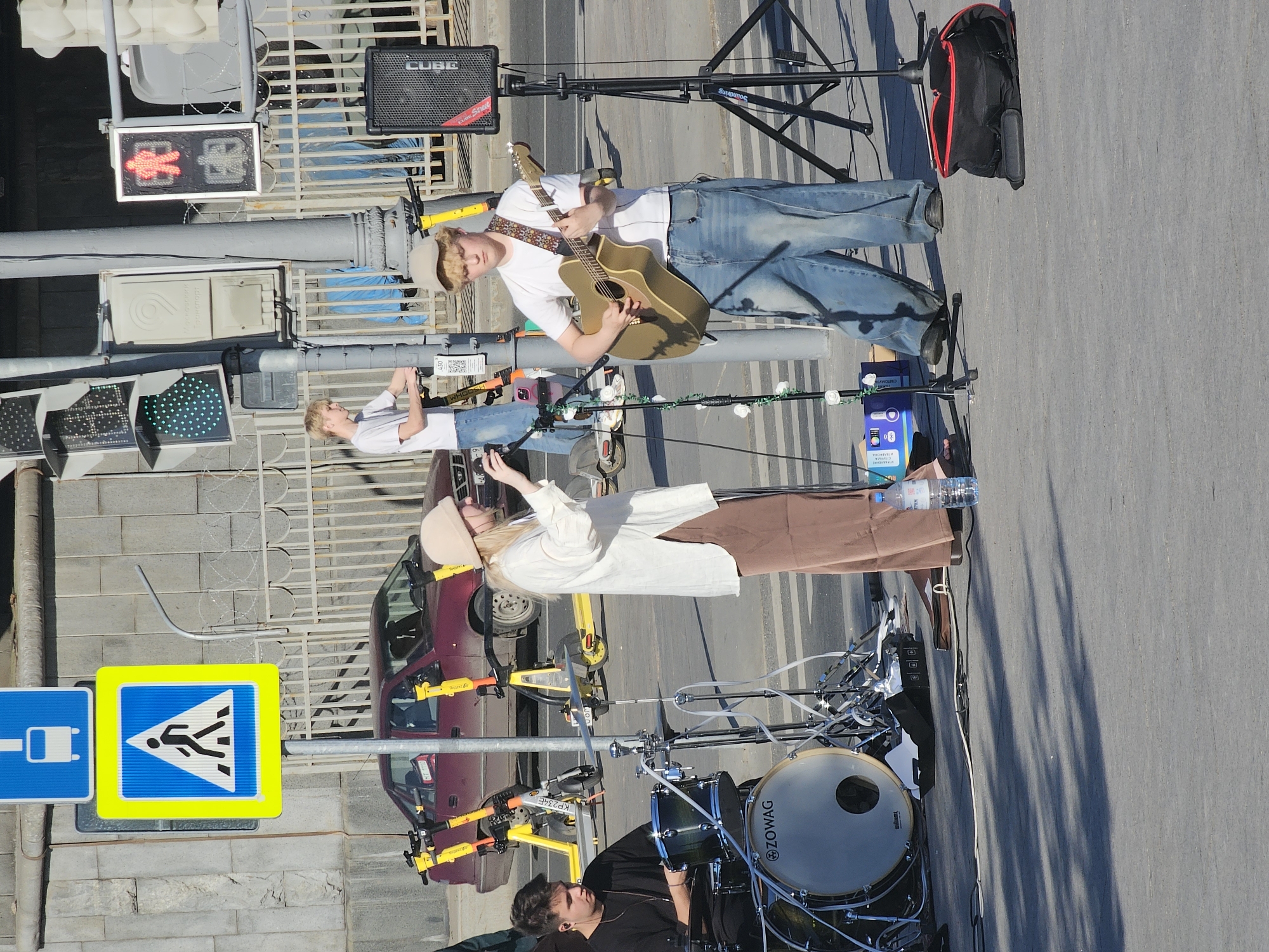 This image captures a street performance taking place on a paved surface in Moscow, Russia. The identification of Moscow is confirmed by a white utility box in the midground labeled "Московский Транспорт" (Moscow Transport) in Cyrillic. In the foreground, a musical group comprising three individuals is performing outdoors. From left to right: 1. A male drummer is seated behind a drum kit, which includes a bass drum featuring the "ZOWAG" logo and a snare drum. He wears a black long-sleeved shirt and has dark hair. 2. A female vocalist or instrumentalist stands centrally, wearing a long-sleeved white tunic-style shirt and wide-legged brown trousers, with a light-colored cap on her head. She holds a small, dark device, possibly a phone or a controller, in her right hand. 3. A male guitarist stands to the right, playing a tan-colored acoustic guitar with a patterned strap. He wears a white short-sleeved t-shirt, baggy blue jeans, and a light-colored baseball cap. Two microphone stands are visible; one to the left of the female performer is adorned with artificial white flowers and green vines, while another plain black stand is positioned to the right of the guitarist, supporting a microphone. A black speaker cabinet with the "CUBE" logo is mounted on a tripod stand to the far right. Various cables, audio equipment, a blue box, and a "ETAN" branded water bottle are scattered on the pavement near the performers. A black bag with red trim rests on the ground to the far right. In the midground, several traffic control elements are present: * A blue rectangular sign depicting a white bus is mounted above a larger yellow square sign with a blue border, featuring a black pedestrian figure walking across a white striped background, indicating a pedestrian crossing. * To the right of these signs are a gray lamppost and multiple traffic light installations. One set of traffic lights displays green circular lights, while another vertical pedestrian traffic light shows a red standing human figure illuminated, with a white walking figure below it, currently unlit. * Behind the traffic elements and performers, a rusty red sedan, possibly a Daewoo Nexia, is parked parallel to a metal fence topped with barbed wire. The partial license plate "HT 110" is visible on the rear of the car. * Numerous yellow electric rental scooters are parked near the lampposts and the red car. * A fourth young male,