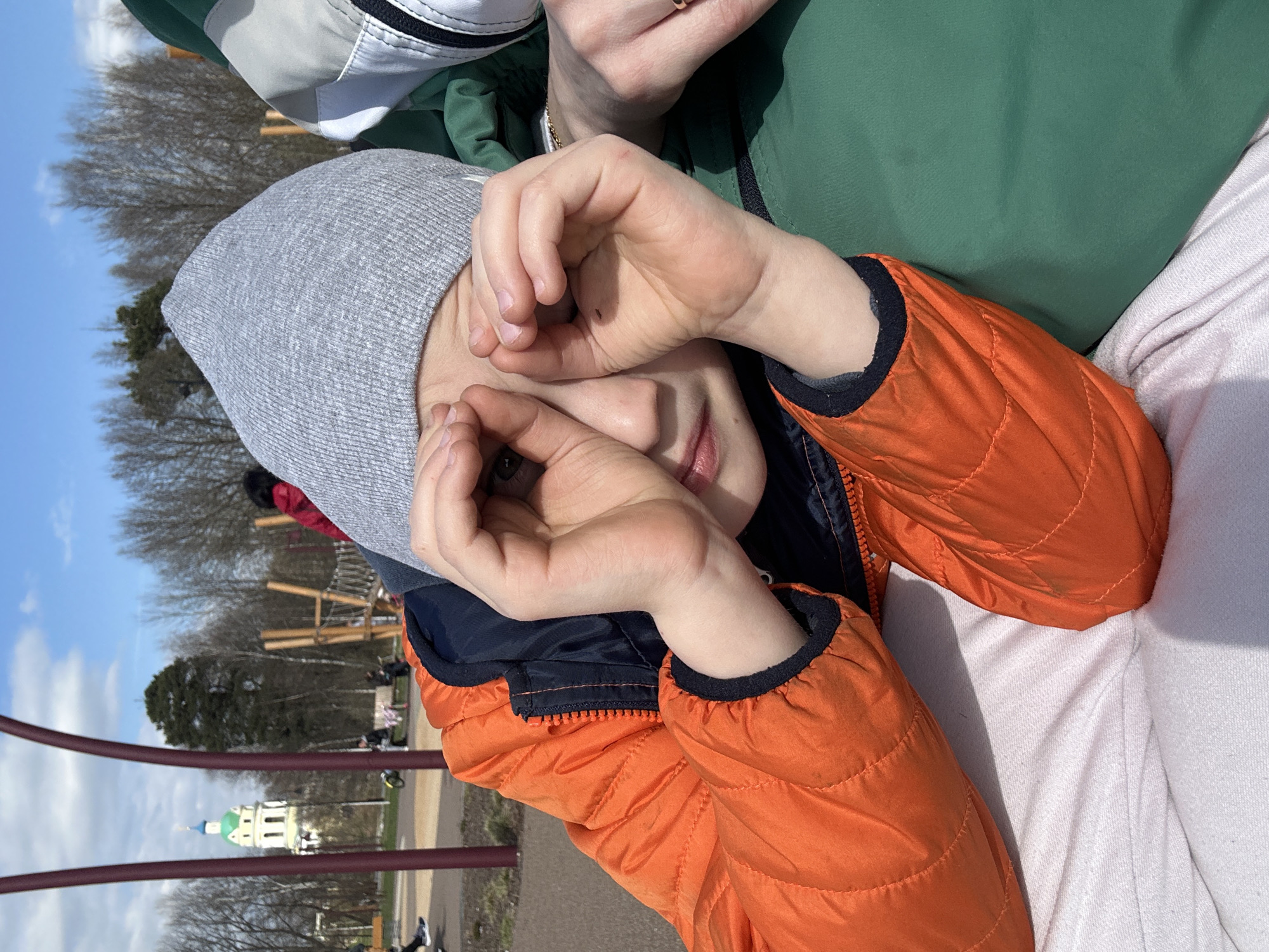 The image captures a child lying down, wearing a gray hat and an orange jacket, with their hands positioned over their eyes. The background reveals a park-like setting, with playground equipment and trees visible. In the distance, the distinctive green and white building of the Church of the Icon of the Mother of God of All Who Sorrow, a landmark in Fryazino, Russia, is identifiable. The overall scene suggests a sunny day in a recreational area.