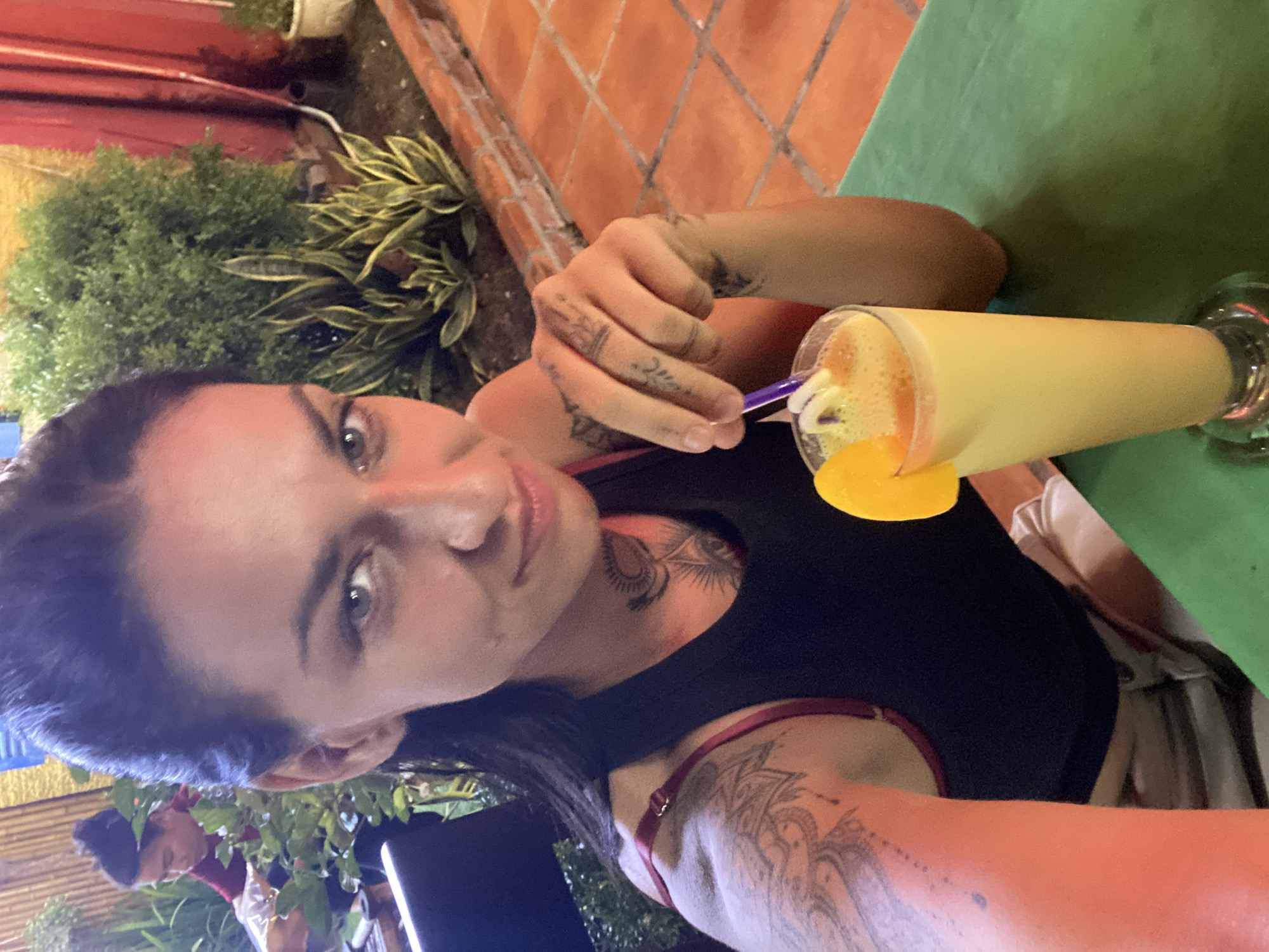 In an outdoor seating area, a woman with dark hair pulled back is taking a selfie. She is wearing a black tank top and has tattoos on her arm, shoulder, and neck. In front of her is a tall glass of a bright orange beverage, garnished with a slice of orange and a straw. To her left, another person with dark hair tied up is visible in the background, wearing a red shirt and tending to some plants. The environment features bamboo fencing, lush greenery, and terracotta tiling on the ground. The lighting suggests it is late afternoon or early evening. The scene is likely in Phu Thuy Ward, Vietnam, given the presence of tropical plants and the overall aesthetic.