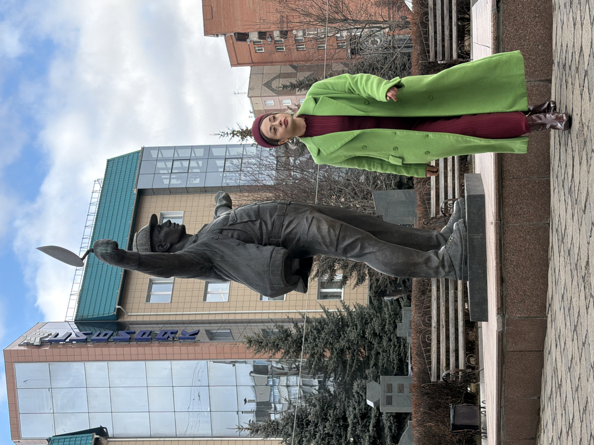 A full-shot outdoor photograph, taken in Krasnoyarsk, Russia, captures a woman standing next to a dark-colored statue of a worker. The scene is an urban public space, likely a plaza or sidewalk, under a partly cloudy sky with patches of blue.
The woman, positioned on the right side of the frame, is dressed in a striking, long, lime green coat, a maroon beret, a dark red or maroon top/dress, and dark brown boots. She stands with her head tilted slightly upwards and to her right, looking into the distance with a neutral expression.
To her left is a statue depicting a male worker, possibly a mason or construction worker, clad in a cap and work clothes. He holds a trowel in his raised right hand, gesturing upwards. The statue, made of a dark material like bronze, stands on a dark rectangular pedestal.
In the background, a modern building features a multi-story glass facade reflecting the sky, topped with a section that has a distinctive green-tiled roof. Above the glass section, blue capital letters are visible, spelling "ПРОЗОРОВ". Further behind, older brick buildings are visible, interspersed with bare trees and evergreen bushes. The ground is paved with patterned grey and brown tiles.
The weather appears cool, consistent with the woman's coat, and the lighting is diffused, suggesting a daytime scene, possibly late morning or early afternoon, with no harsh shadows. The woman seems to be posing for the photograph.