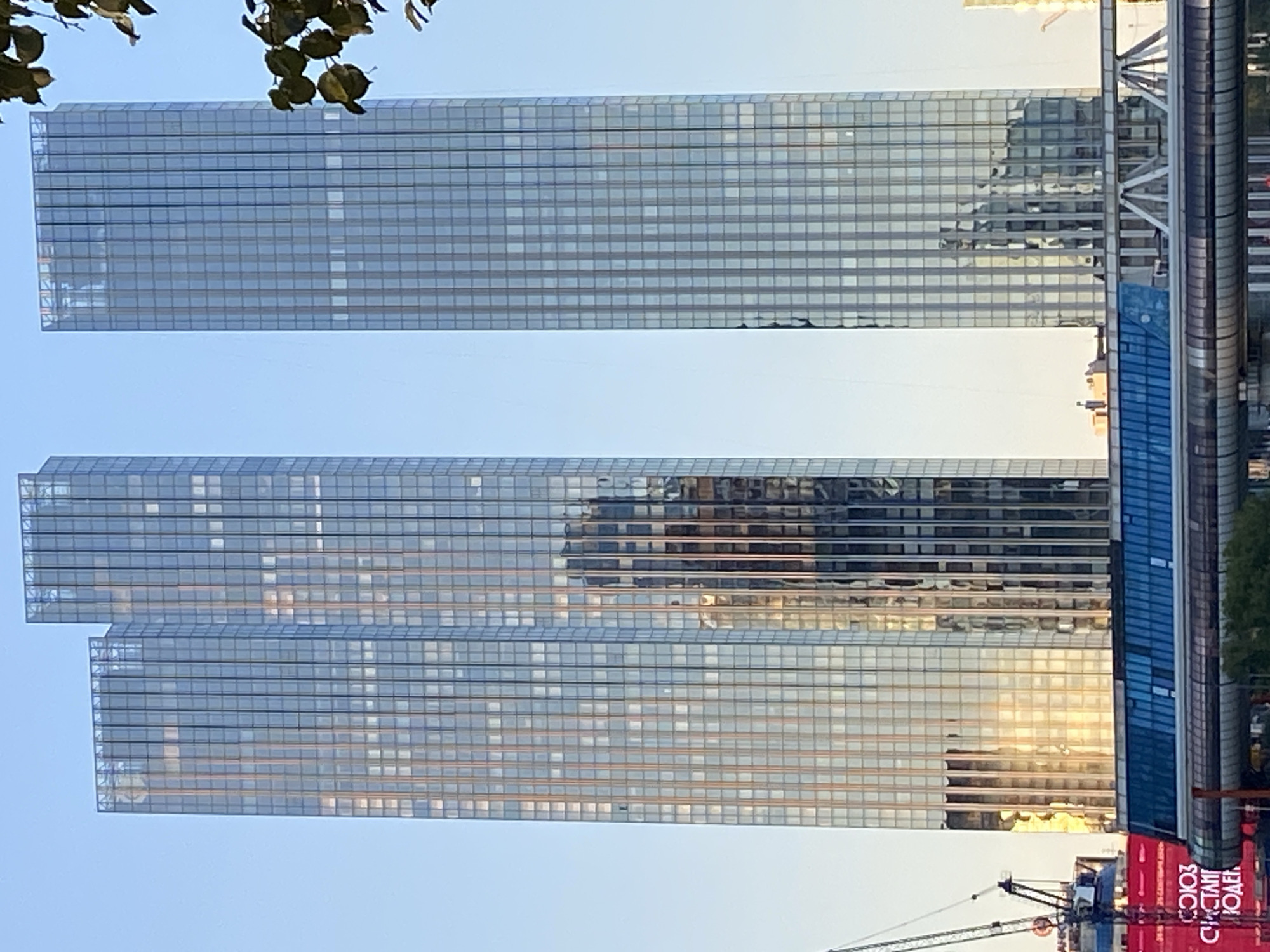 The image shows three modern skyscrapers with glass facades reflecting the clear blue sky and surrounding buildings. The two tallest skyscrapers are very close to each other, with the third one slightly behind and to the left. The facades are composed of numerous rectangular glass panels, with vertical and horizontal lines creating a grid pattern. Sunlight glints off the glass, highlighting some sections and creating reflections. In the foreground, a long, multi-level structure with a blue-tinted glass section and visible support beams spans horizontally. To the left, a construction crane with a red base is visible, and a large red banner with white text, partially obscured by the crane, is present. Some green foliage is seen at the upper right corner of the image, suggesting the presence of trees nearby. The overall impression is of a contemporary urban landscape, possibly a business district.