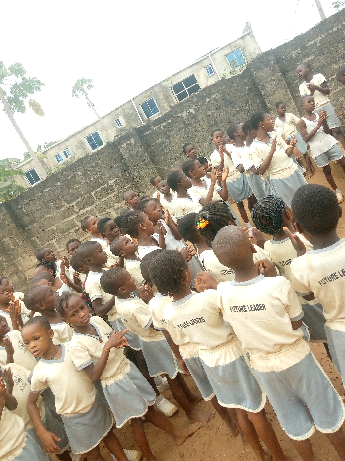 This image depicts an outdoor gathering of approximately 30 children, primarily of primary school age, engaged in an activity in Ukwu-Abwa, Nigeria. The scene is set on unpaved, reddish-brown dirt ground.
**People:** The children are predominantly Black and appear to be between 5 and 10 years old. Most are wearing a uniform consisting of a light-colored (off-white or pale yellow) short-sleeved top and light blue or grey shorts or skirts. Several visible shirts bear the text "FUTURE LEADER" or "FUTURE LEADERS" on the back. Many children are looking upwards and towards the left side of the frame, with their hands raised or appearing to clap. In the foreground, a girl with braided hair and a patterned scrunchie looks towards the camera, pointing her right hand with her index and middle fingers extended. Another girl next to her also looks towards the camera. Most children are standing, some are barefoot, while others wear white shoes.
**Environment:** Behind the children is a weathered, grey concrete block wall, approximately 1.5 to 2 meters high, showing signs of wear and discoloration. Beyond the wall, a multi-story concrete building with multiple windows, some tinted blue, is visible under an overcast sky. Two tall, slender palm-like trees rise in the far left background. The overall lighting suggests an outdoor daytime setting.