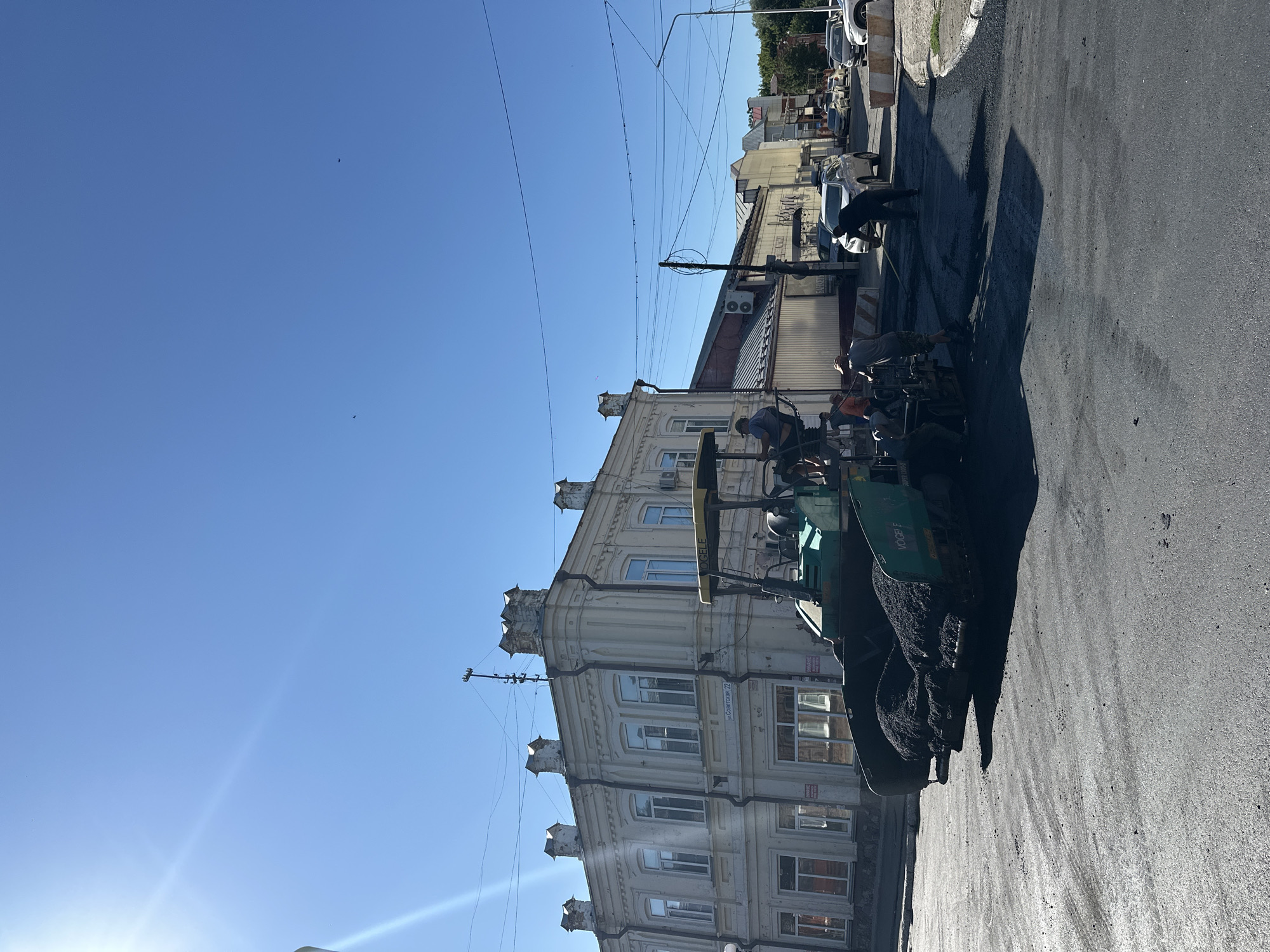 The image depicts road construction in Biysk, Russia, on a clear, sunny day. A paver machine is actively laying down asphalt, with several workers present. One worker is seated on the paver, while others are standing nearby, attending to the machinery and the paving process. The background features a light-colored, ornate building with multiple windows and decorative architectural elements, possibly indicating a historic district or a significant structure. Further down the street, other buildings, parked cars, and utility poles are visible, characteristic of an urban environment. The sky is a vibrant blue, and the sun's rays create a noticeable lens flare effect in the upper left corner of the image. The overall scene captures a moment of ongoing infrastructure work within a city setting.