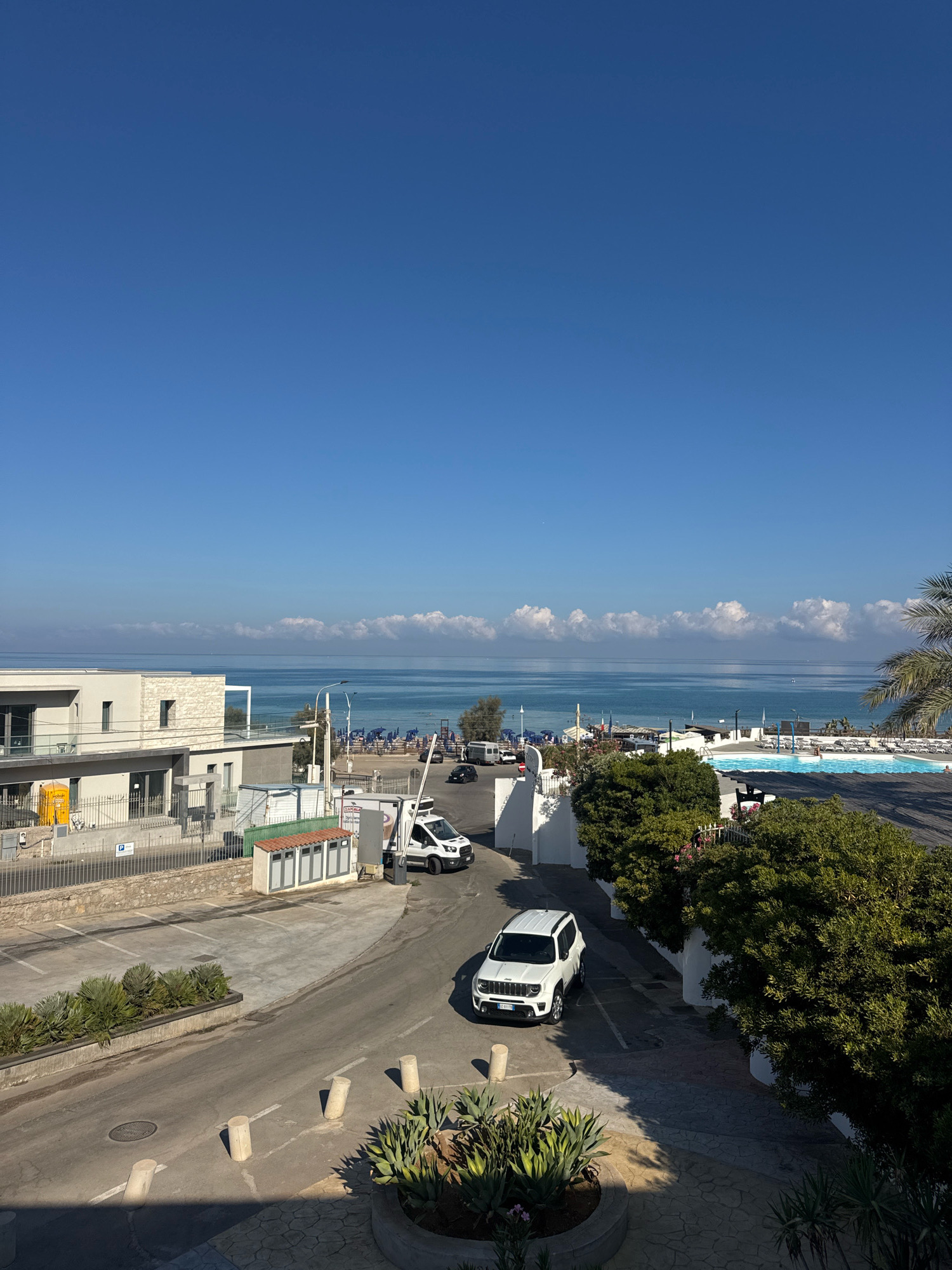 This is a daytime, eye-level, medium shot of a coastal scene in Capaci, Italy. The image captures a view of the Mediterranean Sea, a beach with numerous lounge chairs and umbrellas, and a swimming pool area. On the left side of the frame, there is a modern building with a white facade and balconies. A road runs through the scene, with a white Jeep Renegade parked on it, and a white van at a checkpoint further up the road. There are several trees and plants in the foreground, with shadows cast across the pavement. The sky is clear and blue with a few scattered clouds along the horizon.