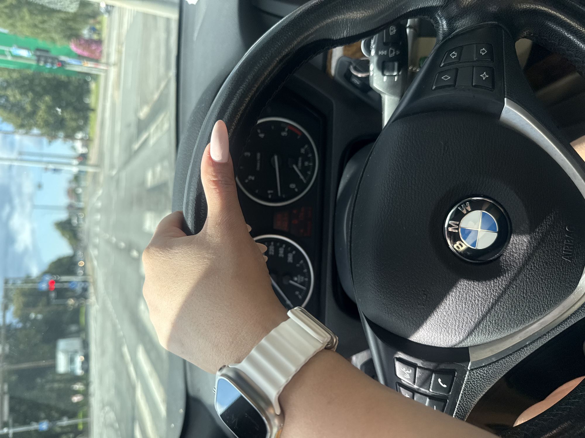 The image shows a person's hand with a white Apple Watch on their wrist, gripping the steering wheel of a BMW vehicle. The steering wheel has buttons on the right side and the BMW logo in the center. The dashboard includes the speedometer and tachometer, with a red traffic light visible in the distance through the windshield. The scene appears to be a street in Tallinn, Estonia, based on the presence of a white truck on the road ahead and trees lining the street. The sky is partly cloudy.
