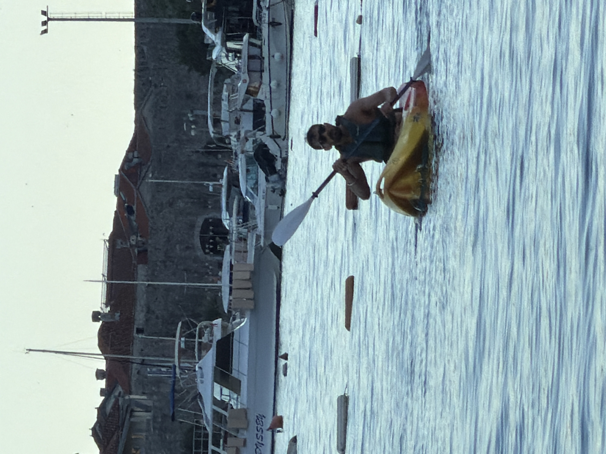 A tanned man with dark hair and sunglasses is paddling a yellow and red kayak on the water. He is wearing a navy blue life vest. He is holding a paddle in both hands. In the background, there are numerous boats docked at the harbor of Budva, Montenegro. The walls of the old town are visible behind the boats.