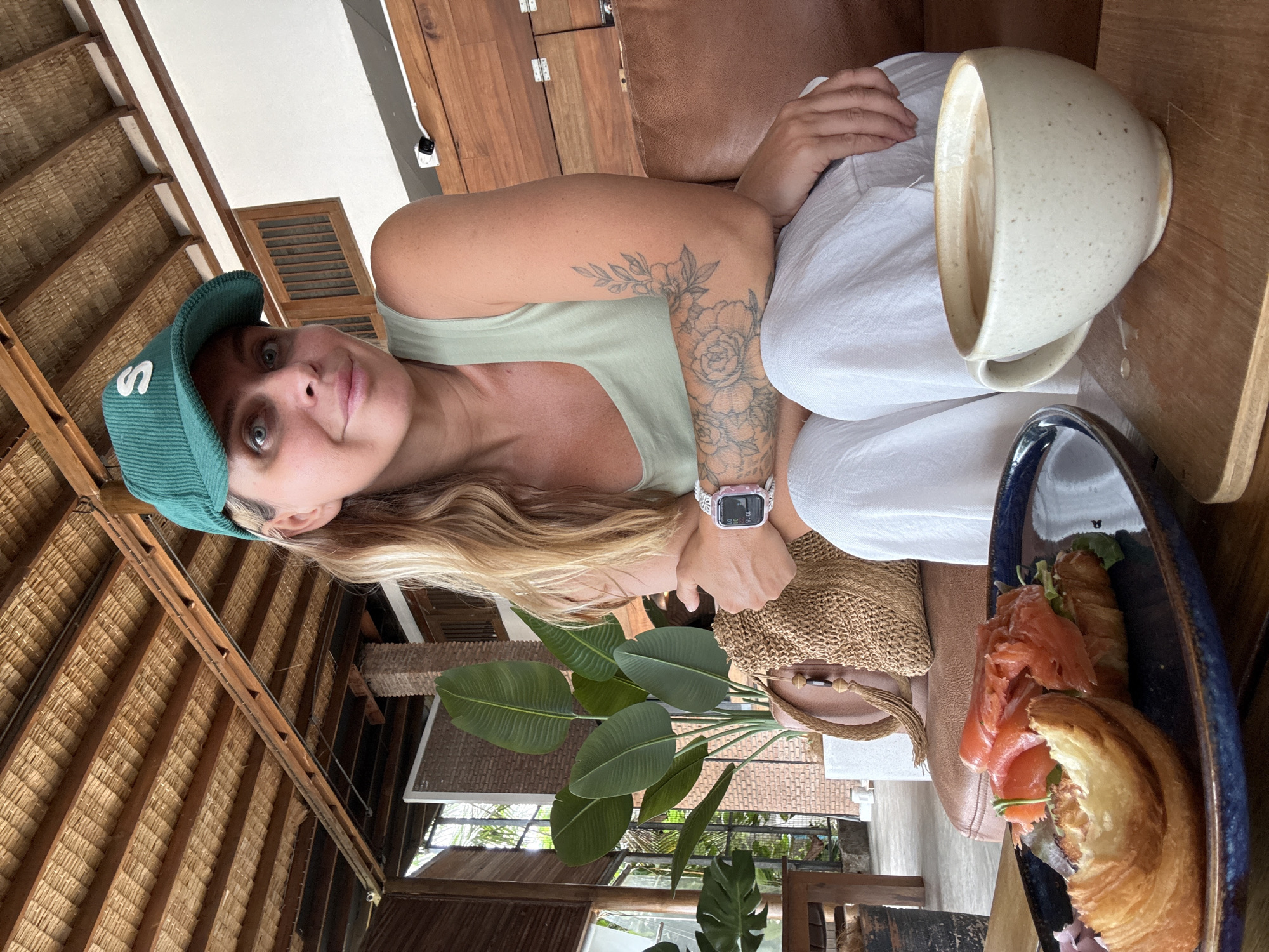 A blonde woman with blue eyes and a green baseball cap is sitting on a brown leather couch in what appears to be a restaurant in Ko Samui District, Thailand. She is wearing a light green tank top and white pants. Her right arm is crossed over her chest, revealing a floral tattoo. In front of her, on a wooden table, is a plate with a croissant topped with smoked salmon and a cup of coffee. Behind her, a large potted plant with broad green leaves is visible, and the ceiling is made of woven reeds and wood.
