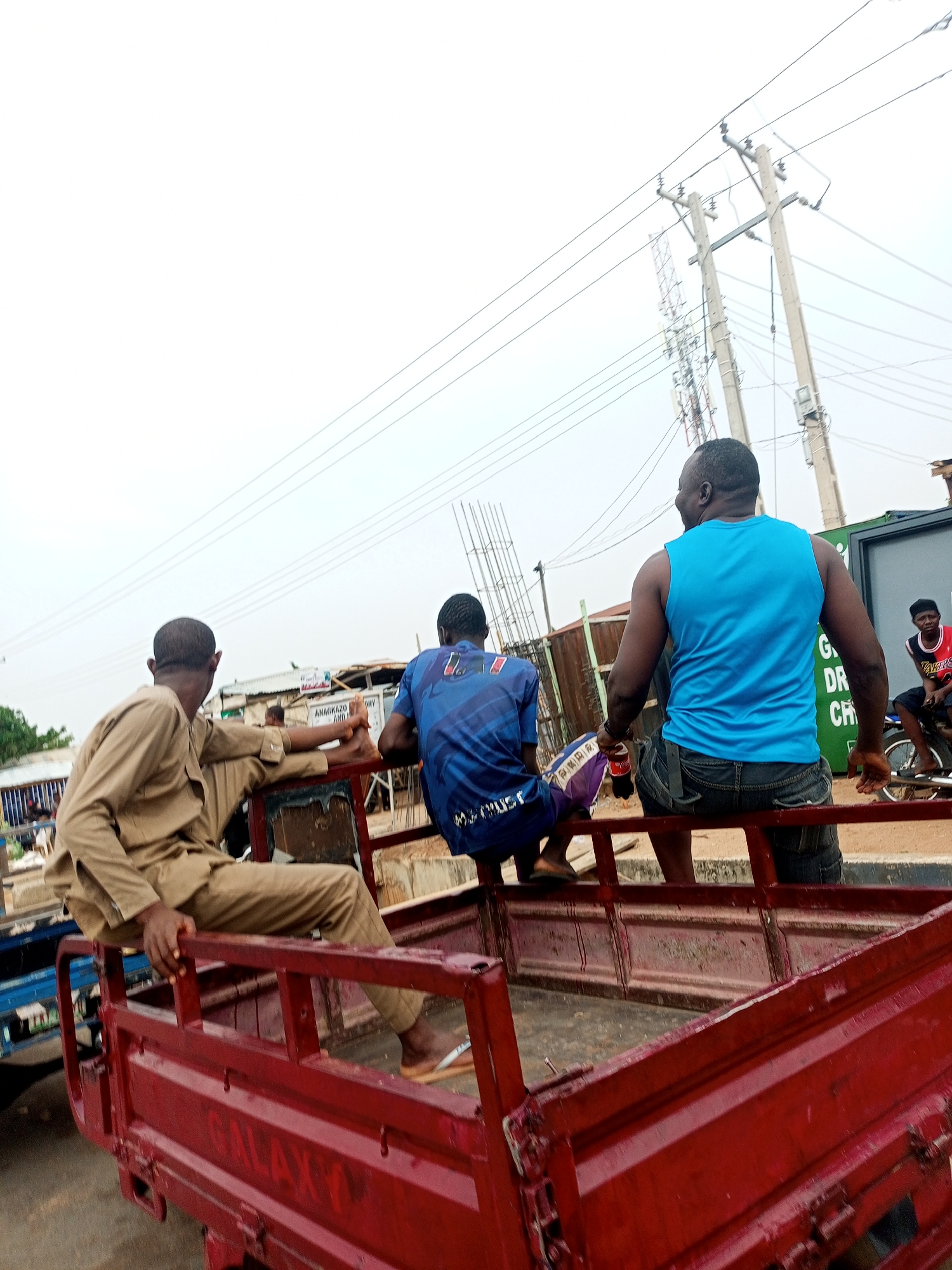 The image captures three men sitting on the back of a red truck in what appears to be a street scene in Tempure, Nigeria. The man on the left wears a tan-colored suit, while the person in the middle is in a blue jersey. The man on the right is wearing a blue tank top and jeans and holding a Coca-Cola bottle. The truck is labeled "GALAXY" on the side. In the background, power lines and poles are visible, along with a green container or structure. Another person wearing a red and black jersey is seated on a motorcycle in the distance.