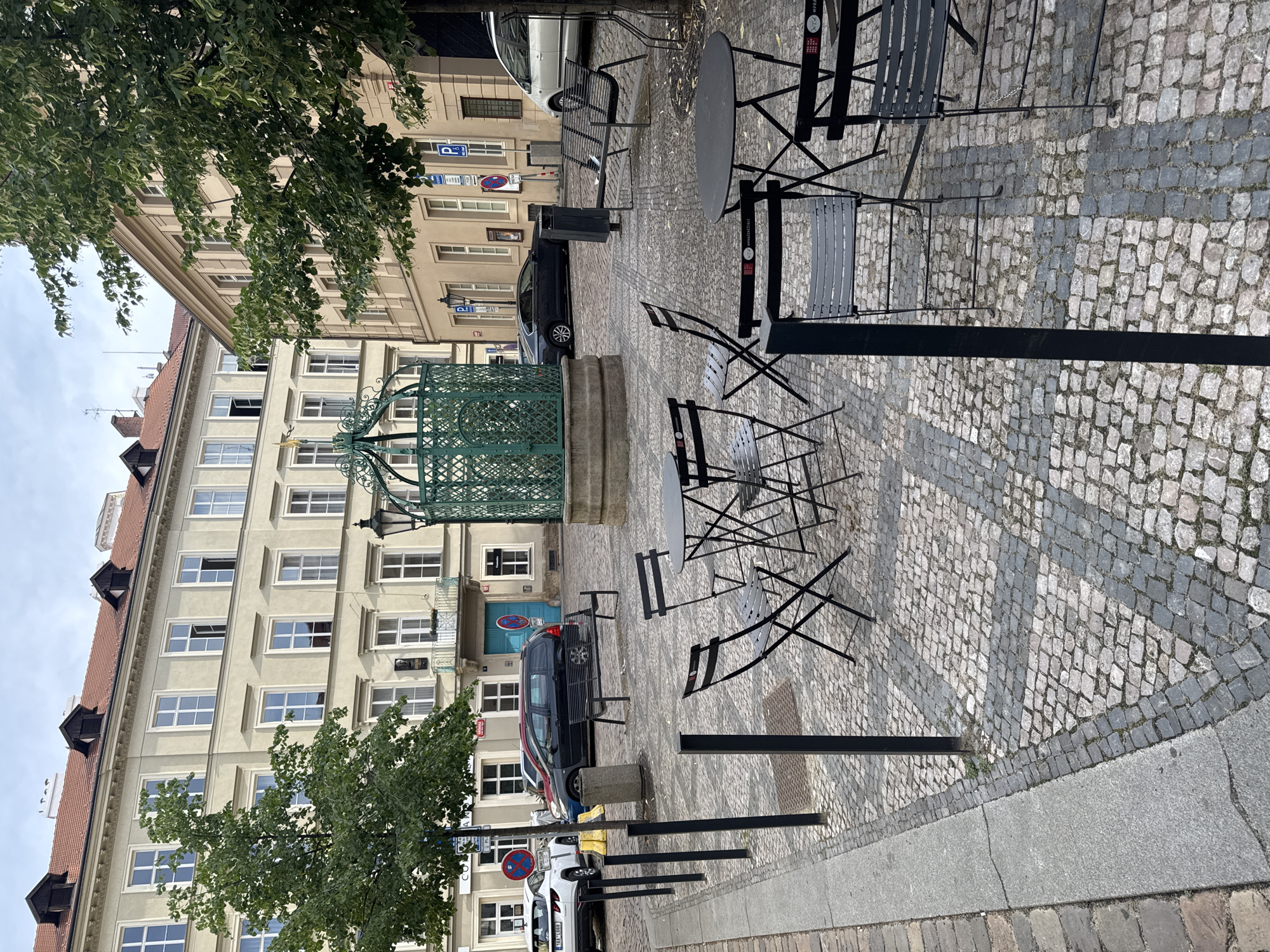 This is a photograph of an outdoor seating area in a city square in Prague, Czechia. The square is paved with cobblestones, and there are several tables and chairs set up, suggesting a cafe or restaurant. In the background, there are buildings with light-colored facades and red tile roofs. One building has an ornate green structure in front of it, possibly a decorative well or a kiosk. There are trees providing shade, and parked cars are visible on the street. The scene appears to be tranquil, with no people present. The lighting suggests it is daytime, possibly overcast.