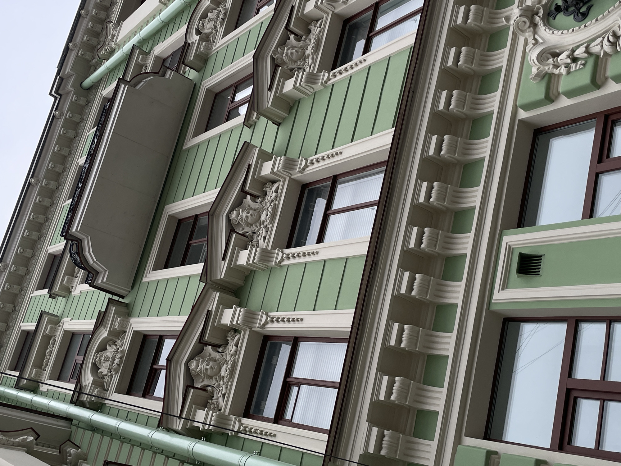 The image shows a section of the exterior of a building in Saint Petersburg, Russia, displaying ornate architectural details and window designs. The building is painted in a light green color with white and brown accents. The facade features decorative moldings, sculpted details resembling faces, and elaborate window frames. The architecture is likely representative of the Art Nouveau or a related style that is prevalent in the historical center of the city. The sky is overcast.