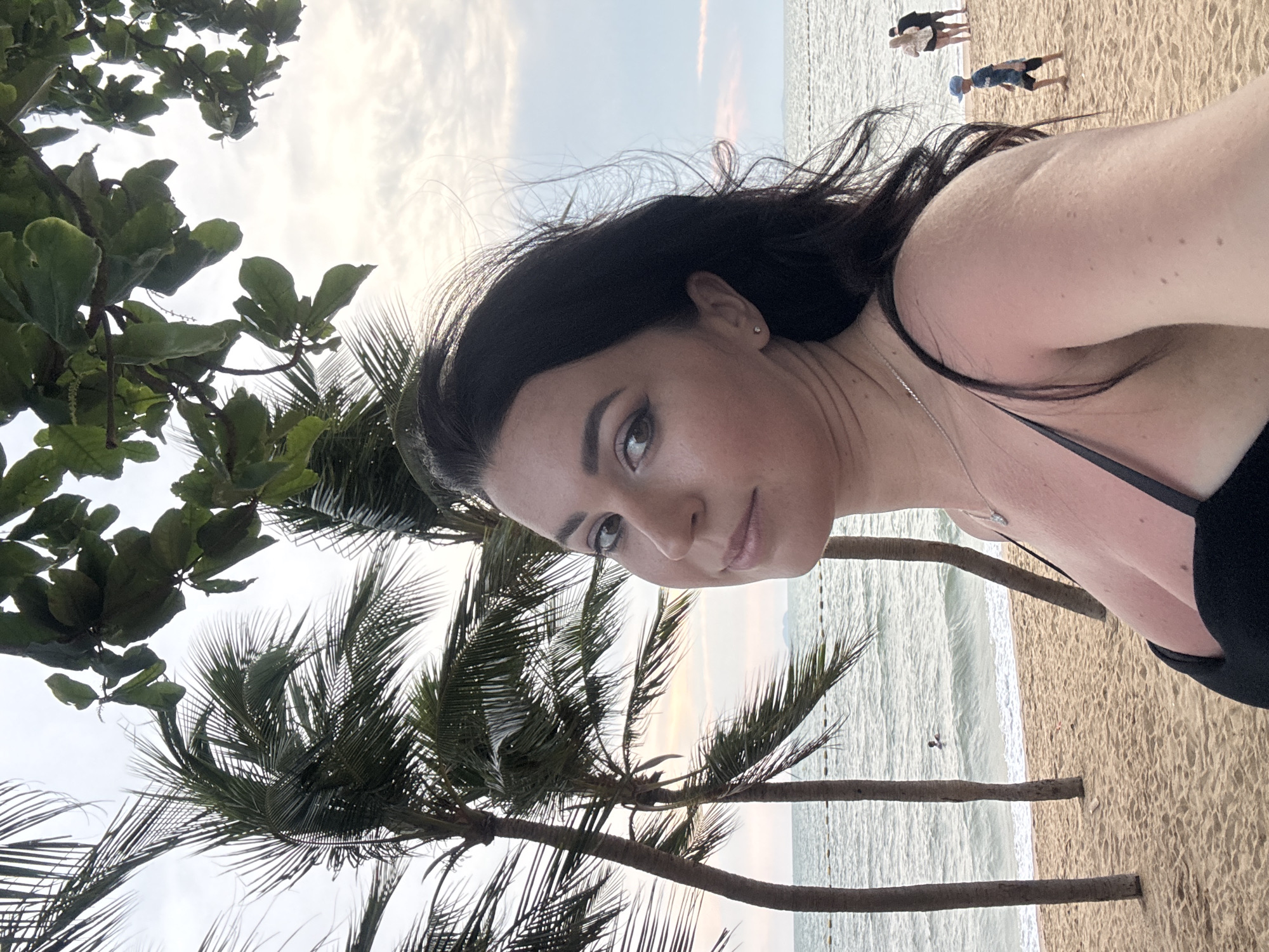 A close-up selfie or portrait shows a woman prominently in the foreground, her face turned slightly to the left with a calm, reflective expression. She has dark, windswept hair, a thin silver necklace, and a small stud earring visible in her right ear, and is wearing a dark-colored top, possibly a swimsuit.
The background features a tropical beach scene. Several large palm trees and other lush green trees frame the shot, their foliage visible against a bright, partly cloudy sky. The ocean stretches across the mid-ground, with gentle waves breaking onto a sandy beach. A buoy line or similar demarcation can be seen in the water.
In the distance on the beach, several small figures are visible; one appears to be a child in a blue hat, and another person stands near the water's edge. The soft, diffused light suggests the late afternoon or early evening, possibly sunset. The setting is a serene, tropical beach environment, consistent with Pattaya City, Thailand. No text is visible in the image.