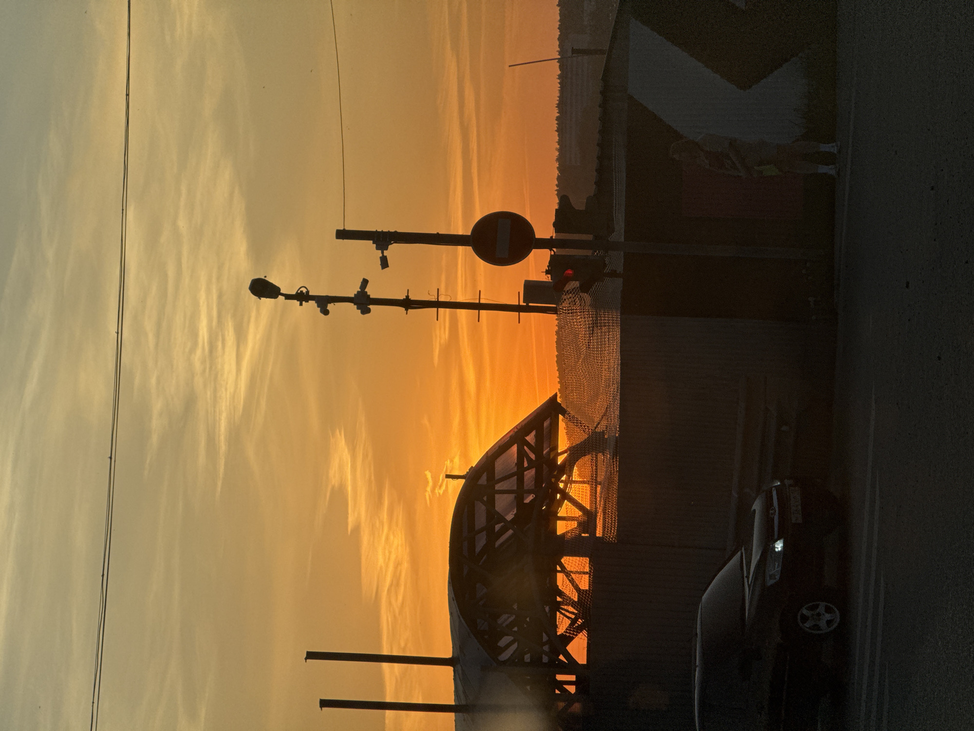 The image depicts a sunset scene with a mix of urban and infrastructure elements. In the foreground, a portion of a dark-colored car is visible on the right side. A metal structure arches across the lower center, possibly a bridge or architectural detail. Behind it, a fence-like barrier stretches across the midground. Several poles rise into the sky, supporting equipment such as security cameras and a "no entry" traffic sign. In the background, a building is partially visible, with a person standing beside it. The sky is filled with orange and yellow hues, indicating a sunset or sunrise. The image captures a moment in a city environment in Izhevsk, Russia.