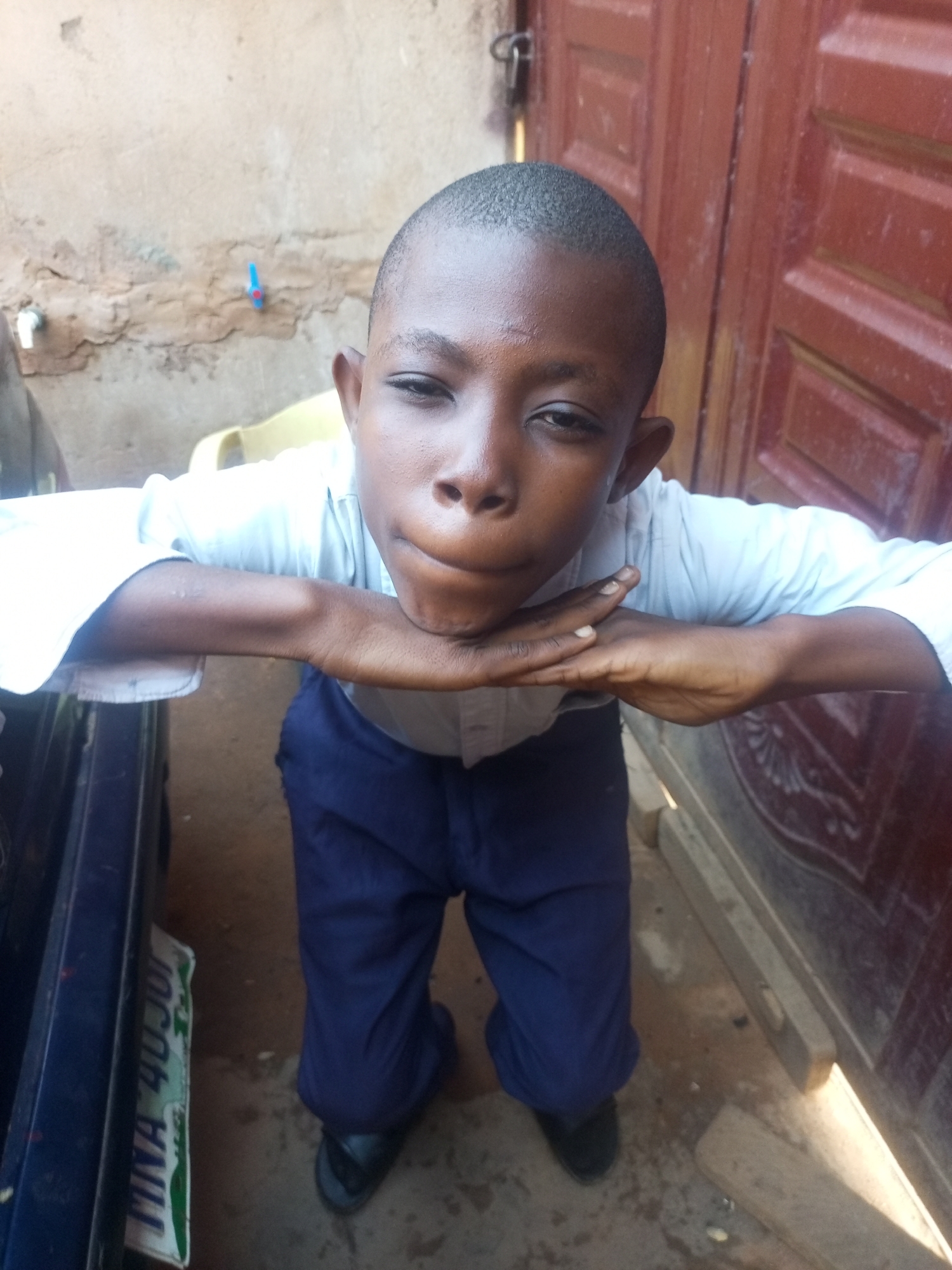 The image shows a young boy standing with his arms resting on his chin, looking directly at the camera. He is wearing a light-colored collared shirt and dark blue pants. His hands are cupped together, supporting his chin. The background consists of a rough, textured wall and a dark red wooden door with vertical panels. To the left of the boy, a portion of a dark blue vehicle is visible, with a license plate that appears to read "MINA 40JUR". The ground is a mix of dirt and concrete. There is a blue faucet on the wall behind the boy. The lighting suggests it is daytime, likely outdoors, possibly in a residential or workshop area in Tempure, Nigeria, given the context.