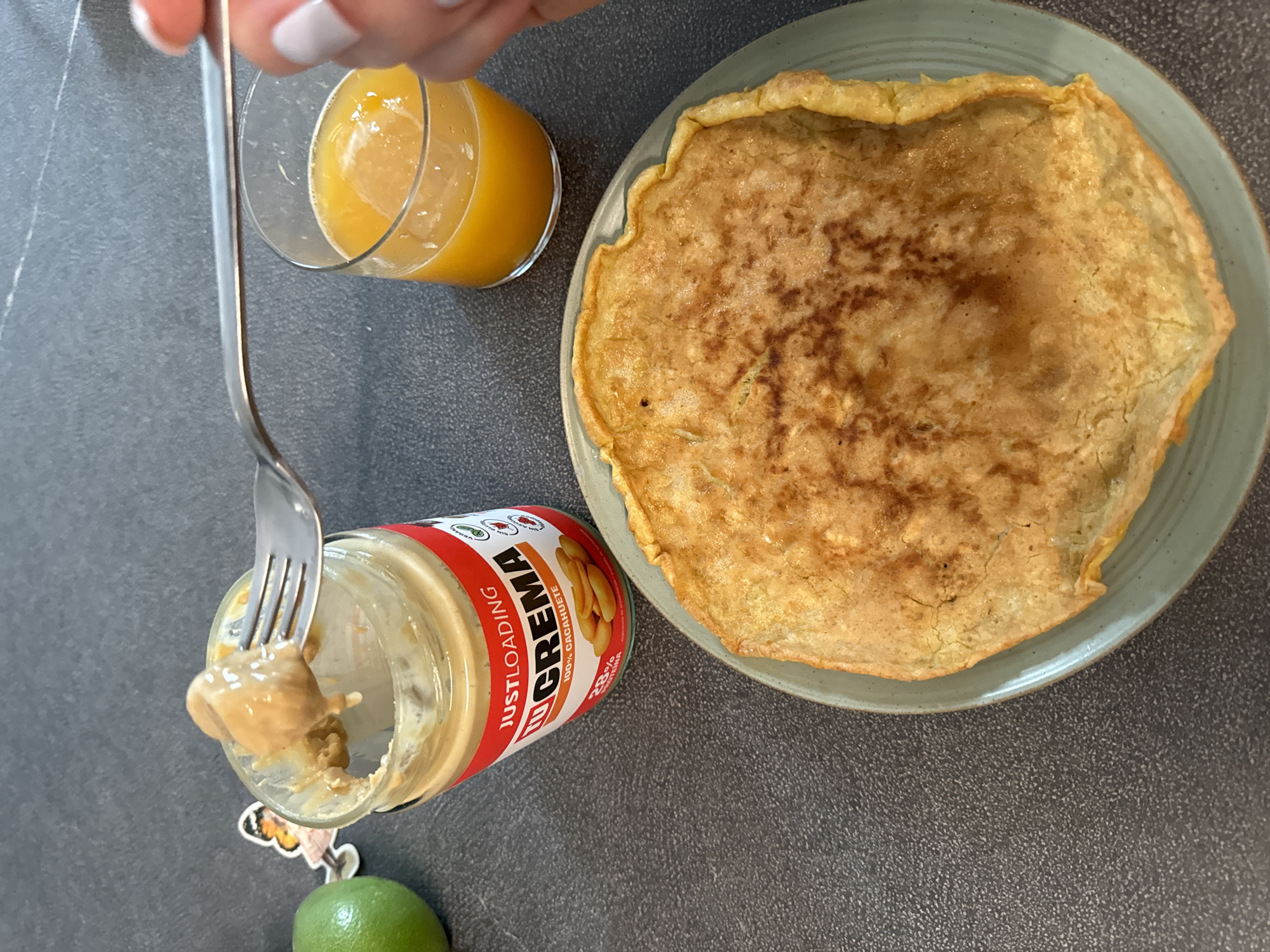This is a close-up, eye-level shot of a breakfast scene on a dark gray, textured countertop. In the foreground, a golden-brown, thin pancake or crepe rests on a light blue-green plate. The pancake has irregular edges and some slightly darker browned spots.
To the left of the pancake, a clear glass filled with a bright orange liquid, likely juice, sits on the counter. A silver fork is held by a person whose hand and manicured nails are visible on the right side of the frame. The fork is in motion, scooping a dollop of creamy, light brown peanut butter from an open jar.
The jar, labeled "JUSTLOADING LU CREMA 100% CACAHUETE," is predominantly white with a red label that features the brand name in bold orange letters. The label also shows images of peanuts and nutritional information like "28% PROTEINA". A small, colorful sticker of a fairy with butterfly wings is attached to the left side of the jar.
To the far left of the frame, a bright green lime is partially visible. The background is the consistent dark gray countertop. There are no architectural elements visible to suggest a specific location within Murcia, Spain. The focus is on the food and ingredients.
