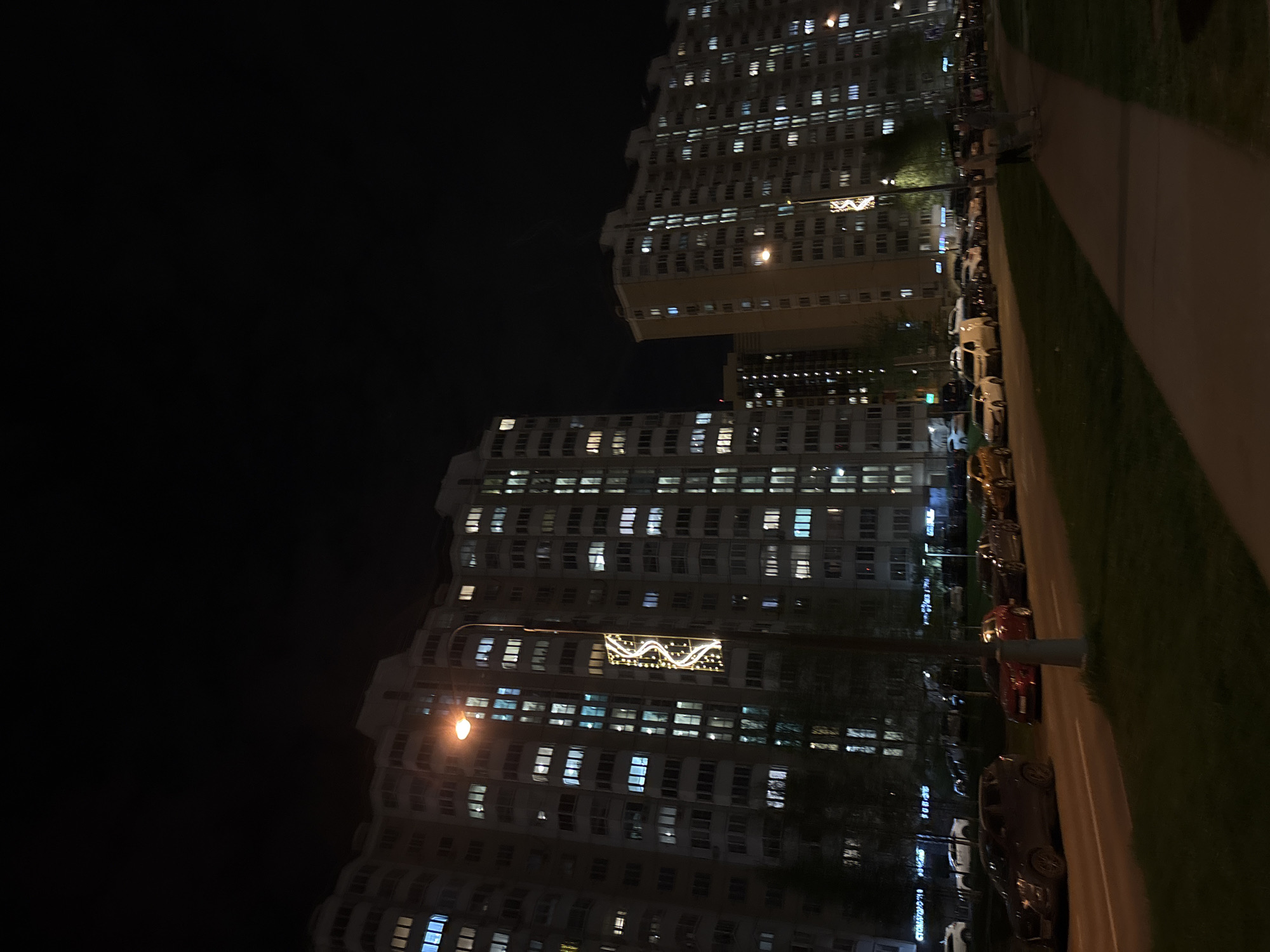 At night, the city of Vnukovo, Russia, is lit by illuminated apartment buildings. A streetlamp provides light for a sidewalk next to a strip of grass. Parked cars line the curb.